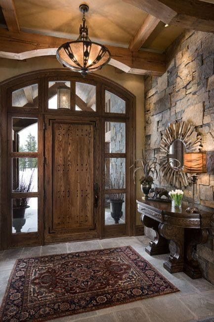 Elegant entryway with a wooden front door and large glass side panels, stone accent walls, a decorative mirror, a small console table with a lamp and plants, a patterned rug, and a chandelier hanging from a wood-beamed ceiling.