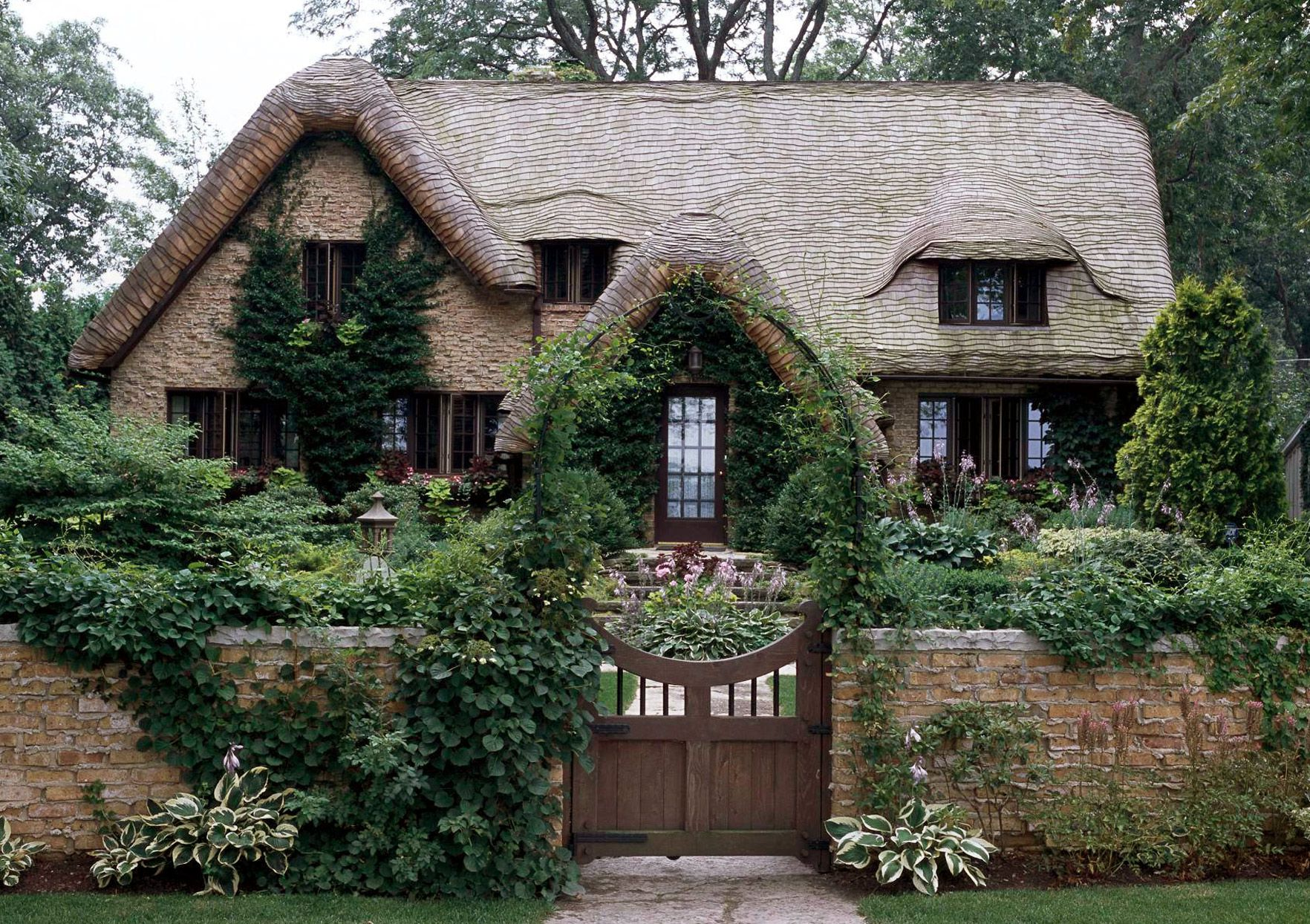 A charming house with a stone exterior and a uniquely shaped, tiled roof. The house is surrounded by lush green plants and trees, with a small garden and a wooden gate at the front.