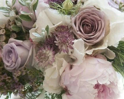 Close-up of a bouquet of pale purple roses, white peonies, and purple flowers with green foliage.