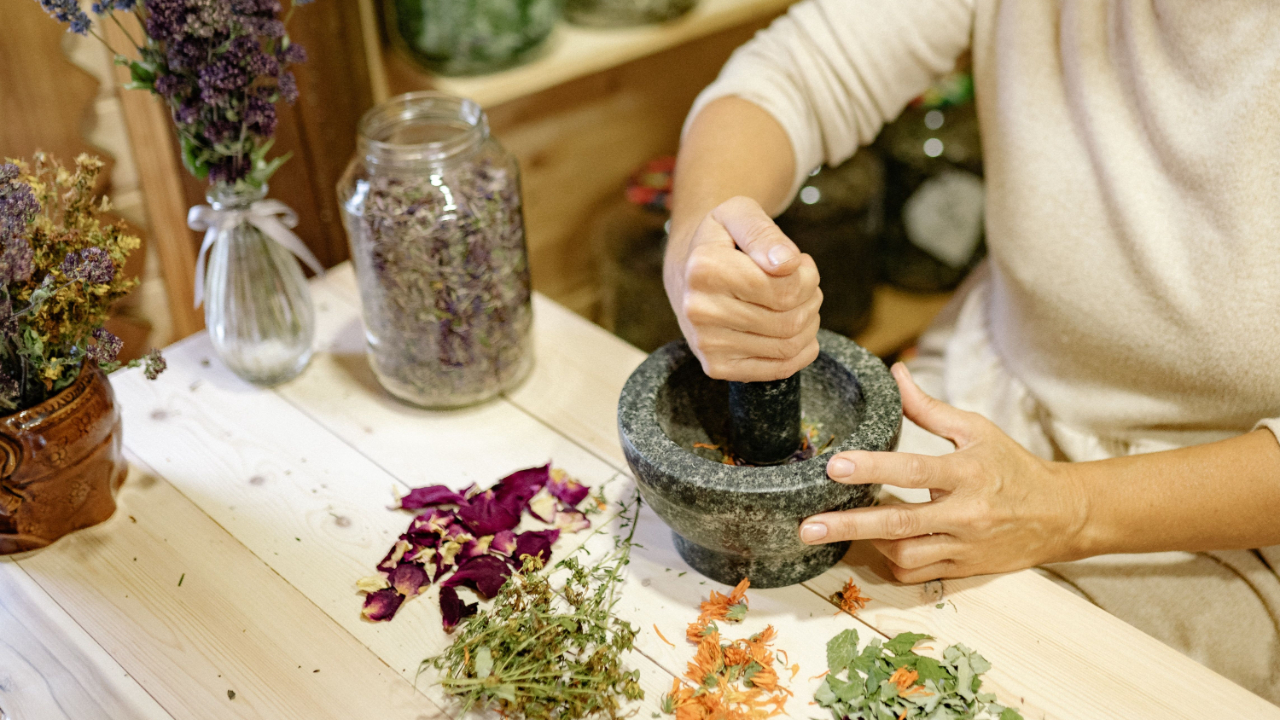 Person using a mortar and pestle to crush dried herbs or flowers on a wooden table with various dried plants and flowers around.