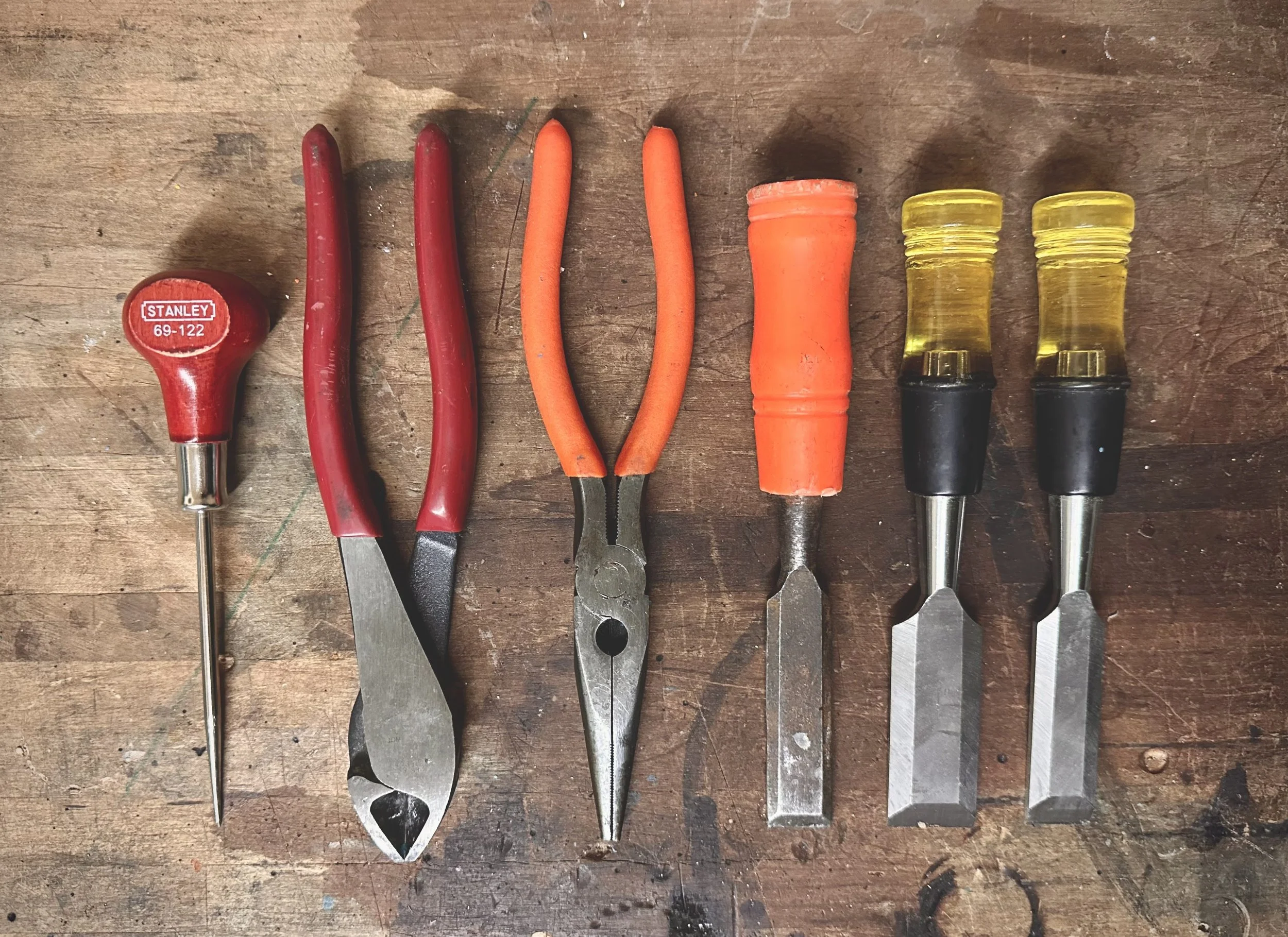 A set of hand tools, including a red-handled awl, two pair of pliers with red and orange handles, a chisel with an orange handle, a flathead screwdriver with a yellow handle, and a Phillips screwdriver with a yellow handle, laid out on a wooden workbench.