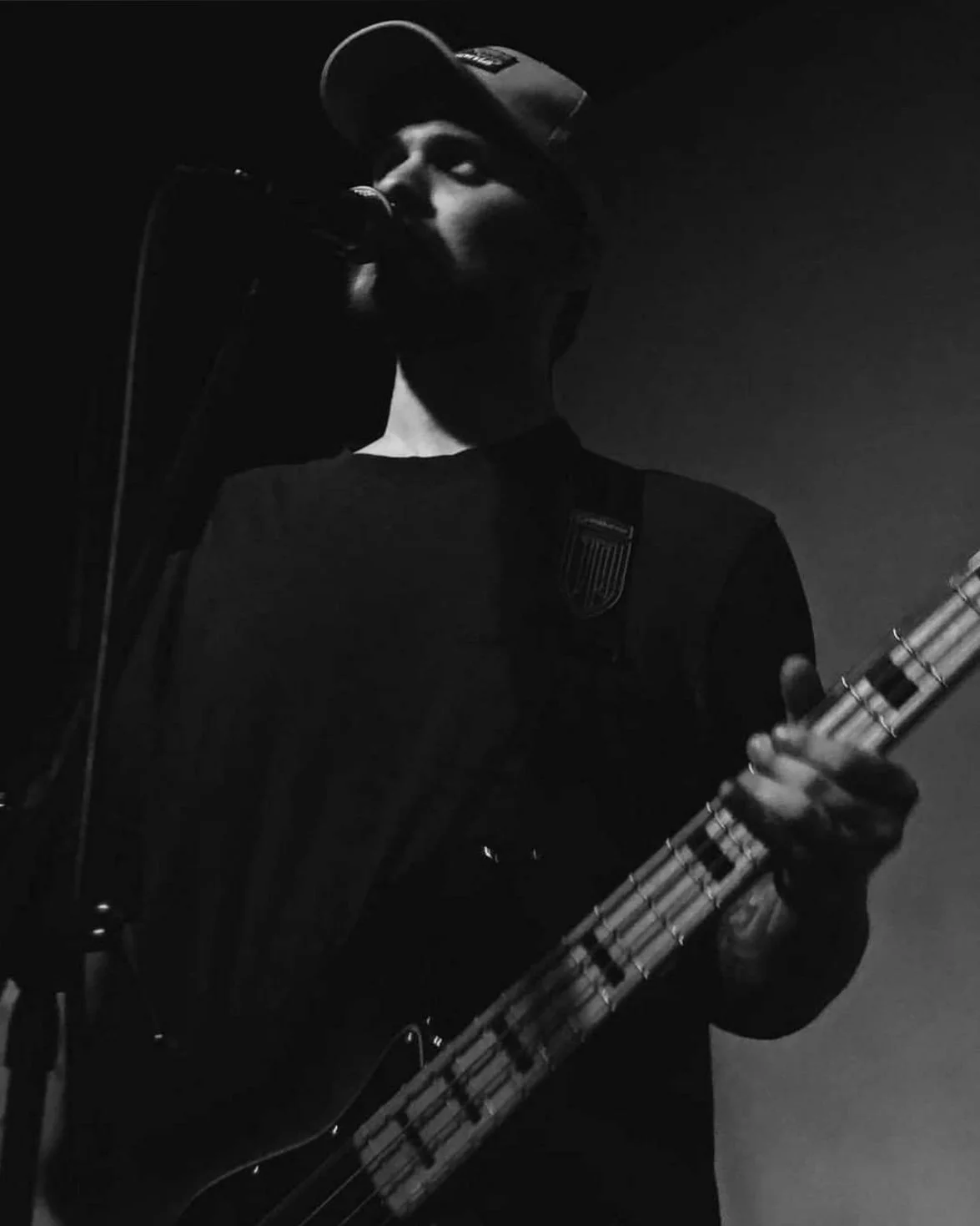 Black and white photo of a person wearing a cap, singing into a microphone, and playing an electric bass guitar.
