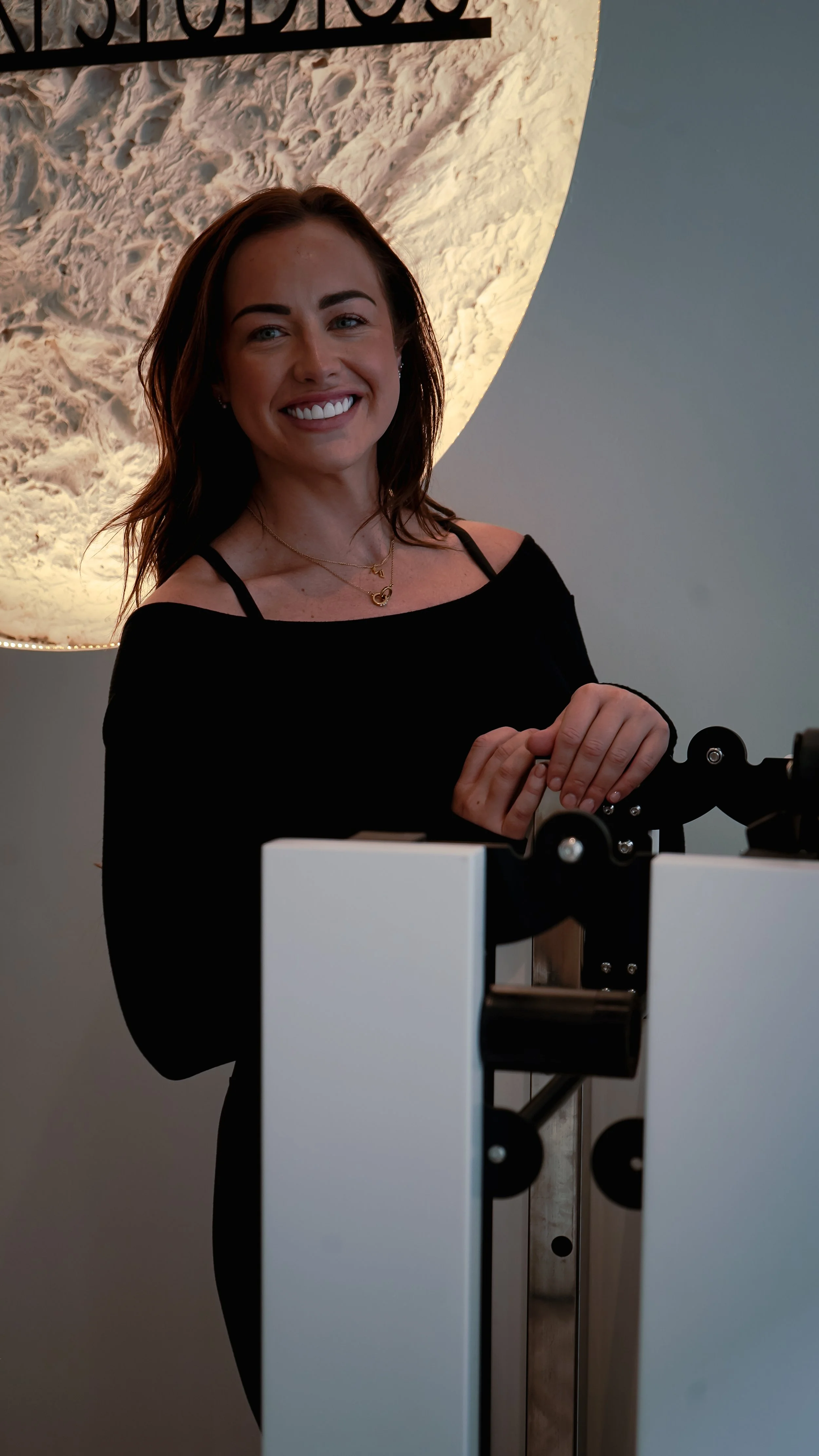 A woman with shoulder-length brown hair smiling, wearing a black off-shoulder top and layered necklaces, standing indoors in front of a moon-like textured wall or artwork.