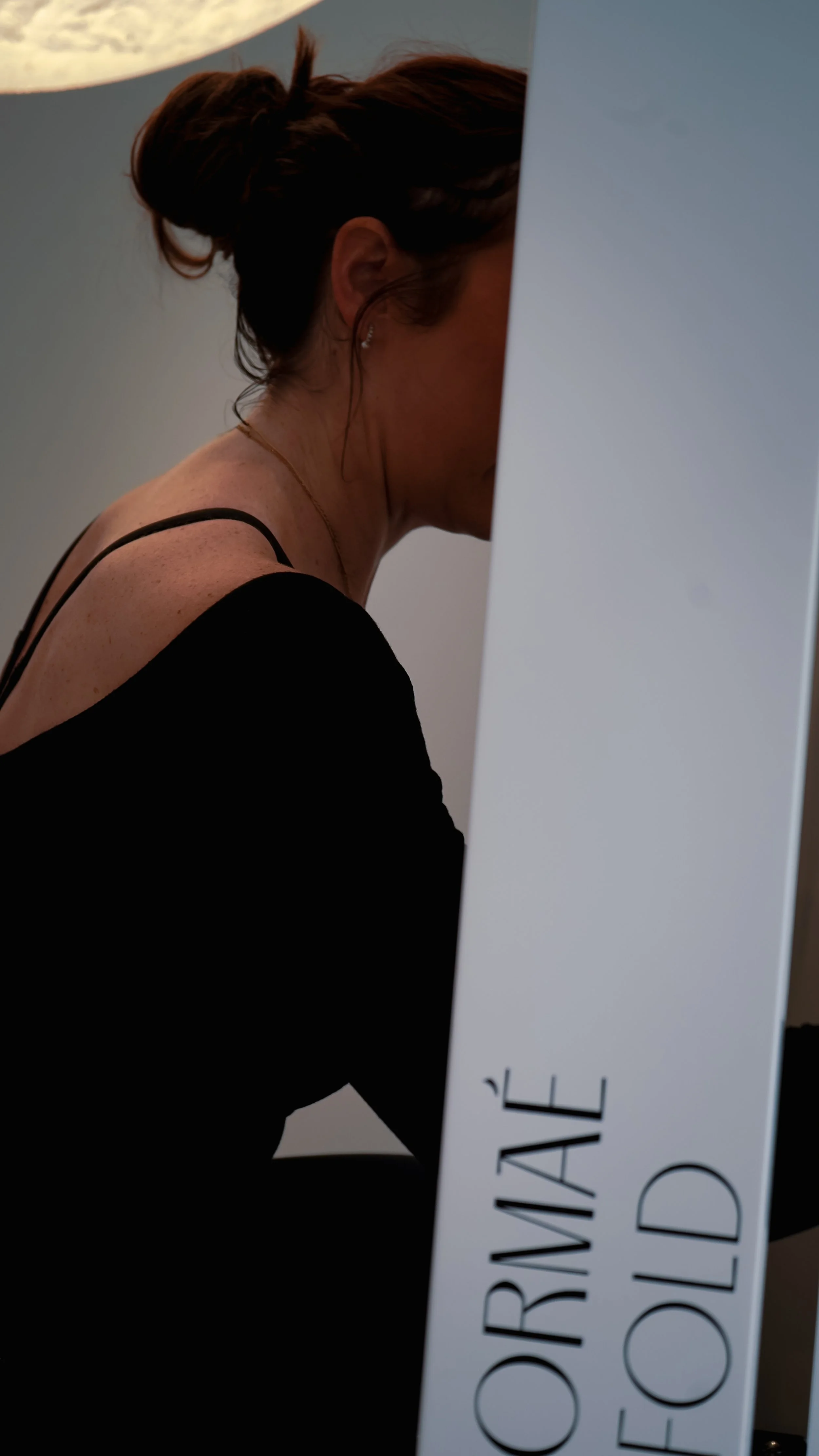 A woman with brown hair tied in a bun, wearing a black top, is seated with her face turned down and partially covered by a large white sign that reads 'ORIGINAL' and 'OF'.