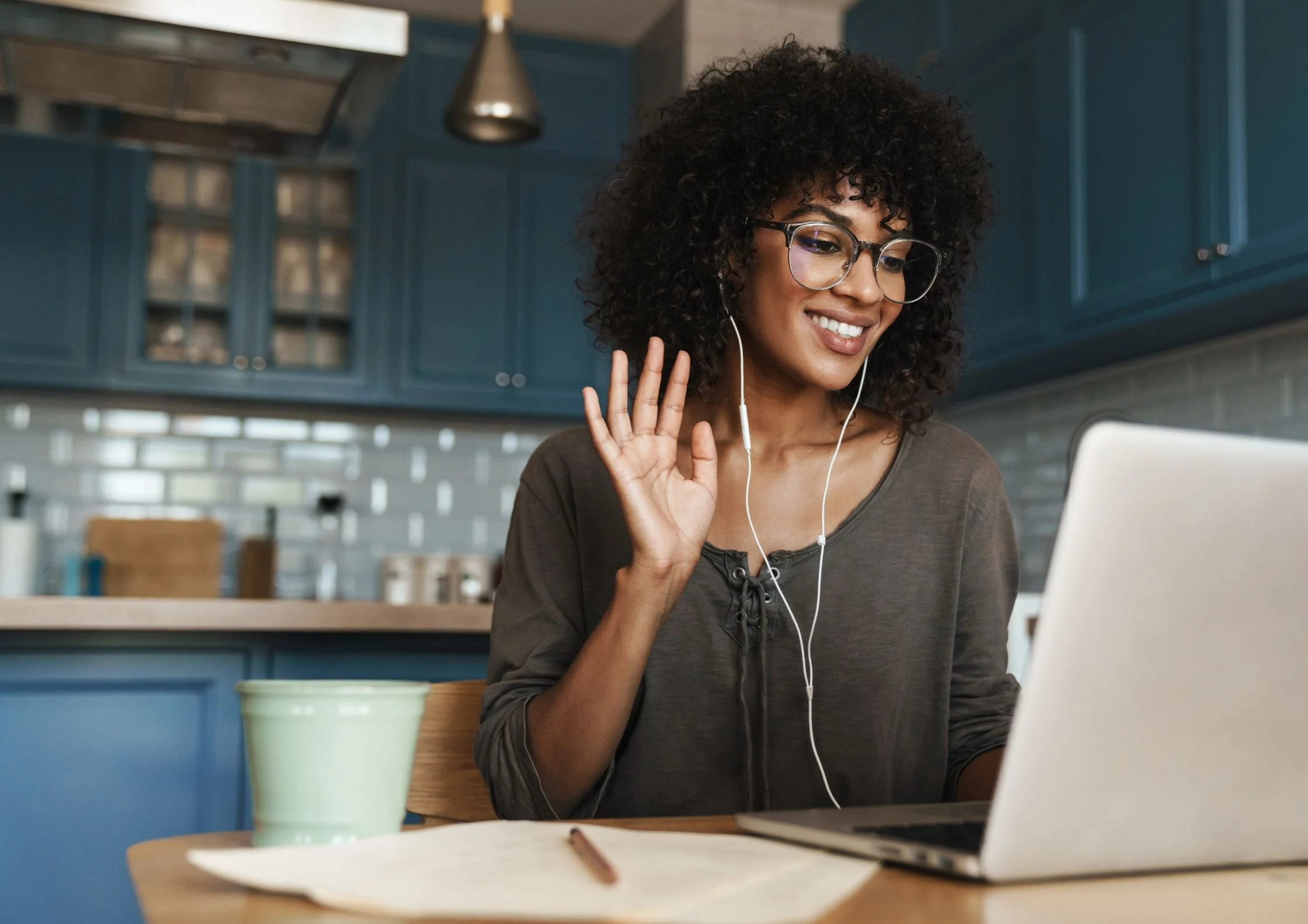 Young woman with curly hair and glasses, smiling and waving while looking at her laptop in a kitchen.