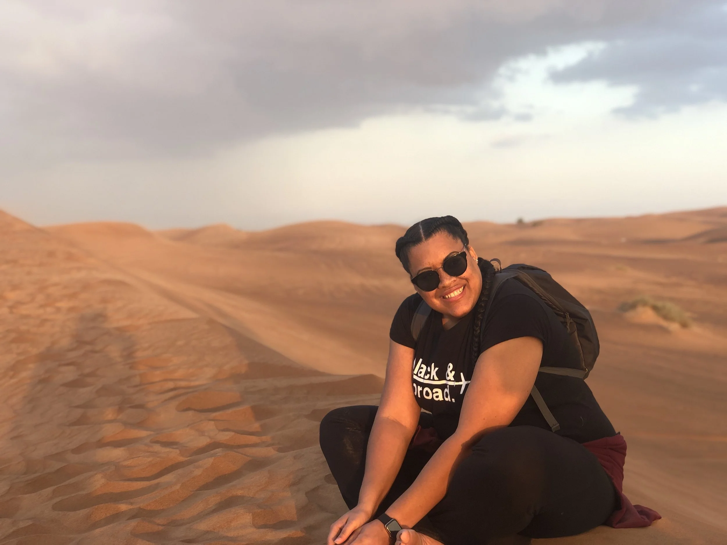 A woman with dark hair in braids, wearing sunglasses, a black t-shirt, and black pants, sitting on sandy dunes with a backpack, smiling at the camera, with a cloudy sky in the background during sunset.