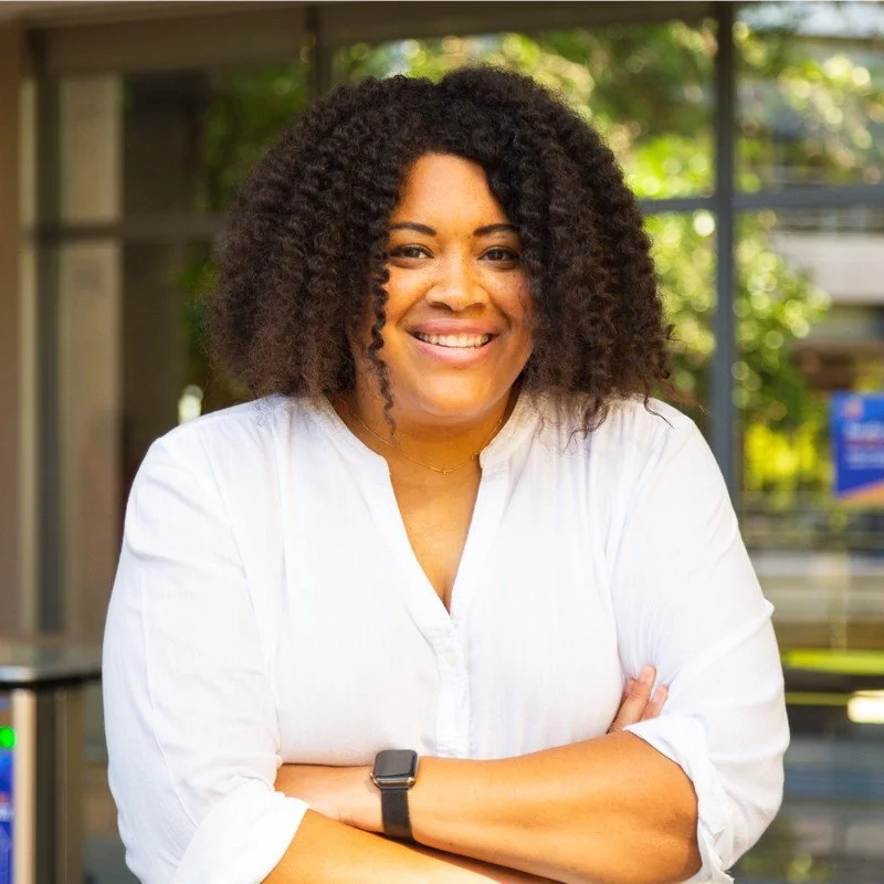 A smiling woman with curly hair wearing a white shirt, standing outdoors with a blurred background of green trees and glass windows.
