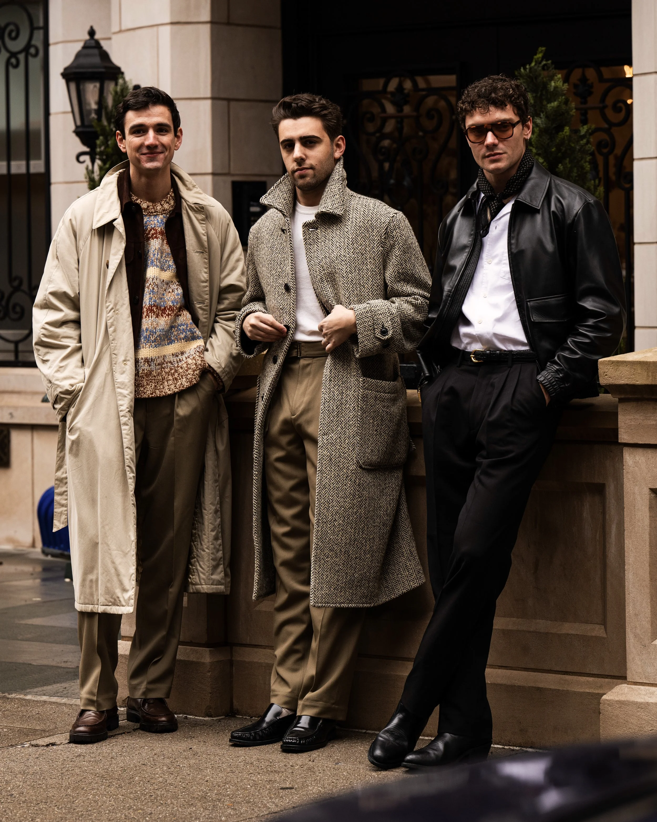 Three men in stylish 1980s fashion standing outside a building with stone walls and black wrought iron fencing. Street style photograph by street style photographer Kyle Mark Peterson during NYFW.