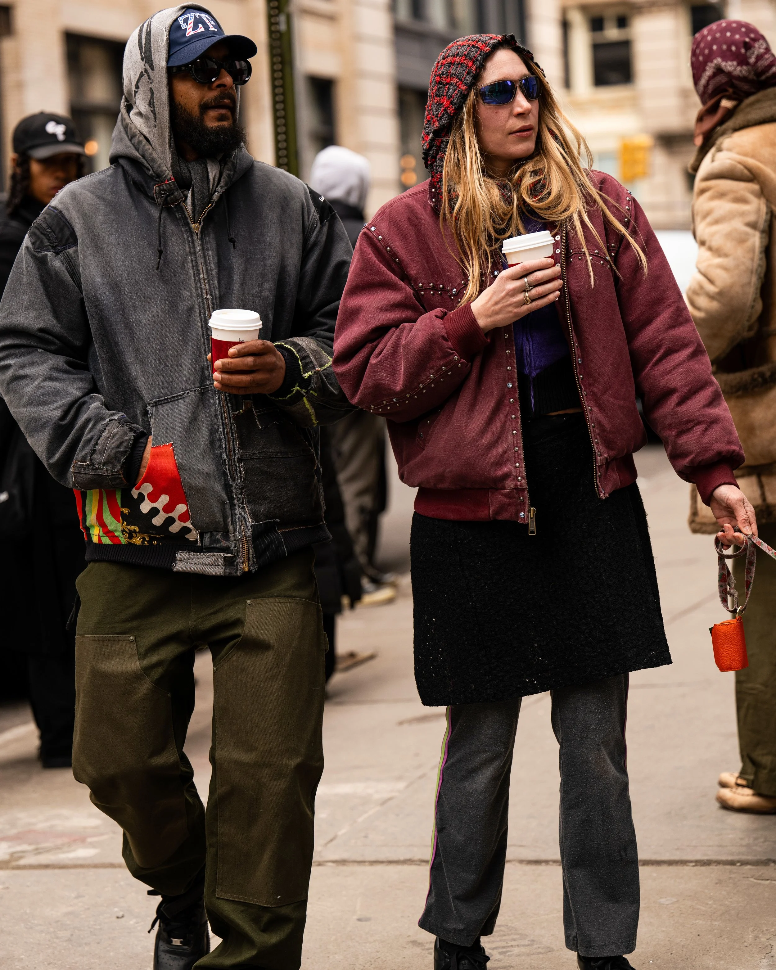Two people walking on city sidewalk holding coffee cups, dressed warmly with jackets and sunglasses.