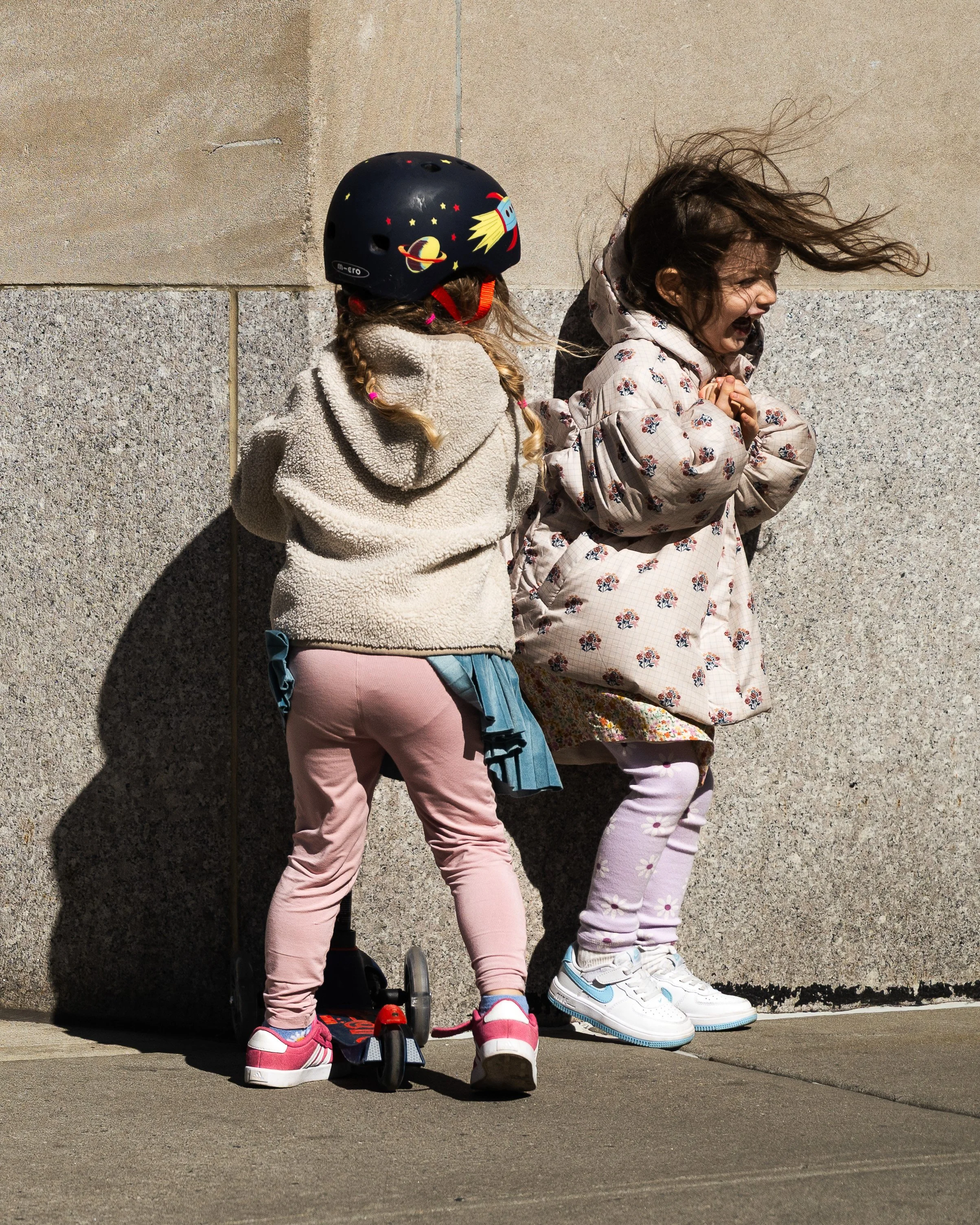 Two young girls are standing against a stone wall. One girl is wearing a helmet with space-themed designs, a beige jacket, pink pants, and pink shoes, riding a scooter. The other girl is wearing a floral coat and light purple pants, and she appears distressed or upset.