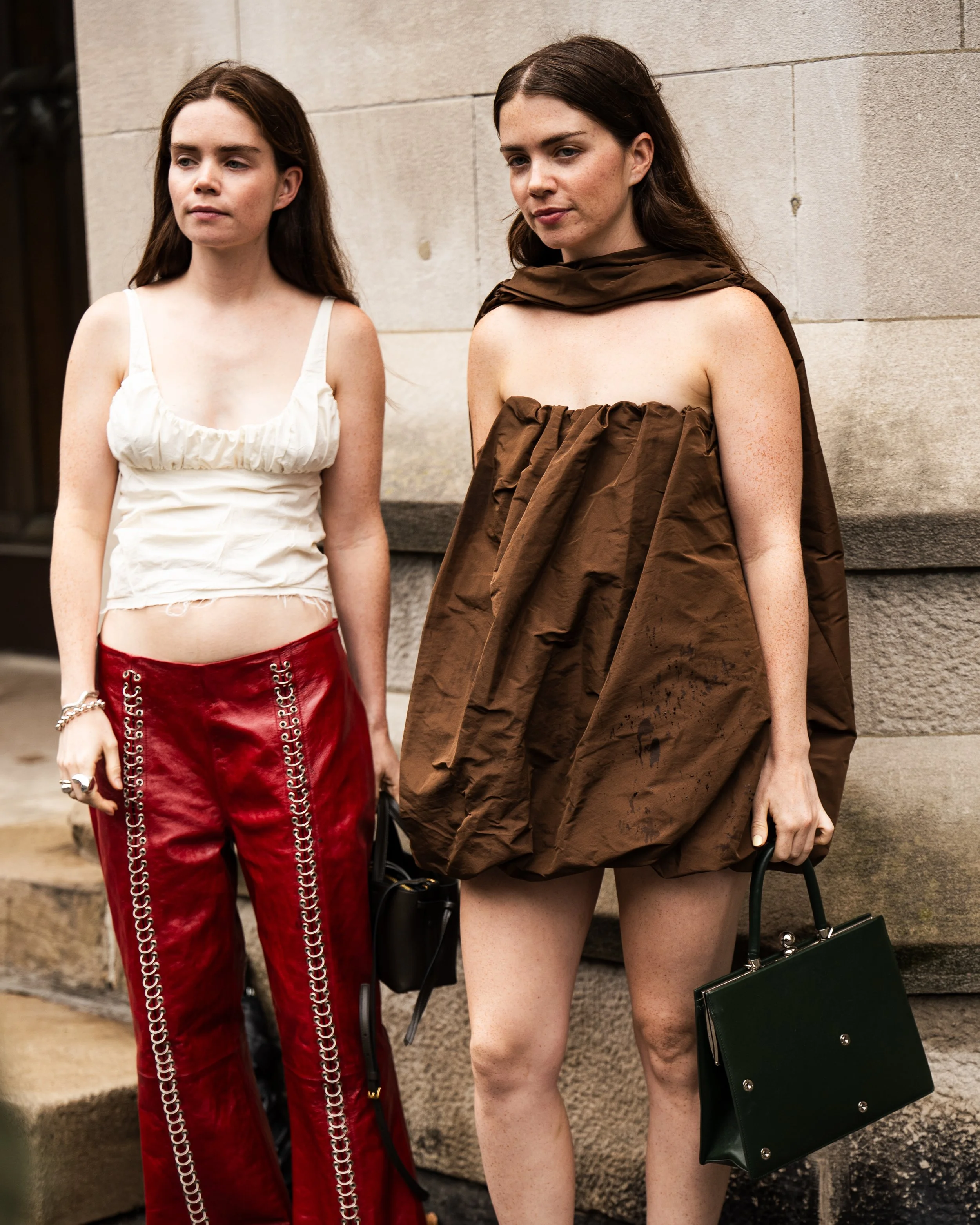 Two women standing against a stone wall, one wearing a white top and red pants with metal chains, the other wearing a brown dress made of crinkled fabric, holding a black handbag. Street style photograph by street style photographer. NYFW.