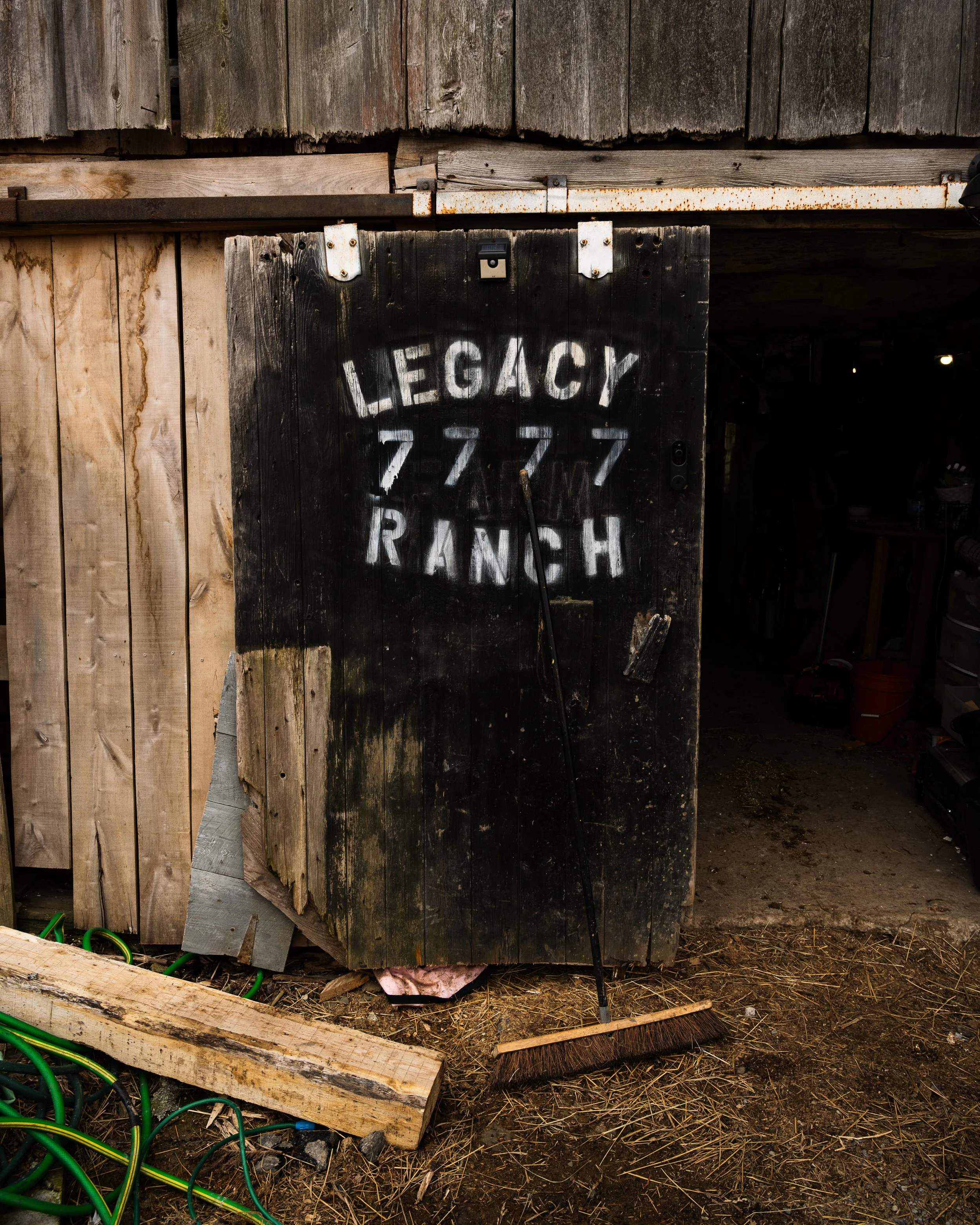 Old wooden barn door painted with white letters reading 'Legacy 777 Ranch' on a dark background, with a broom leaning against it.