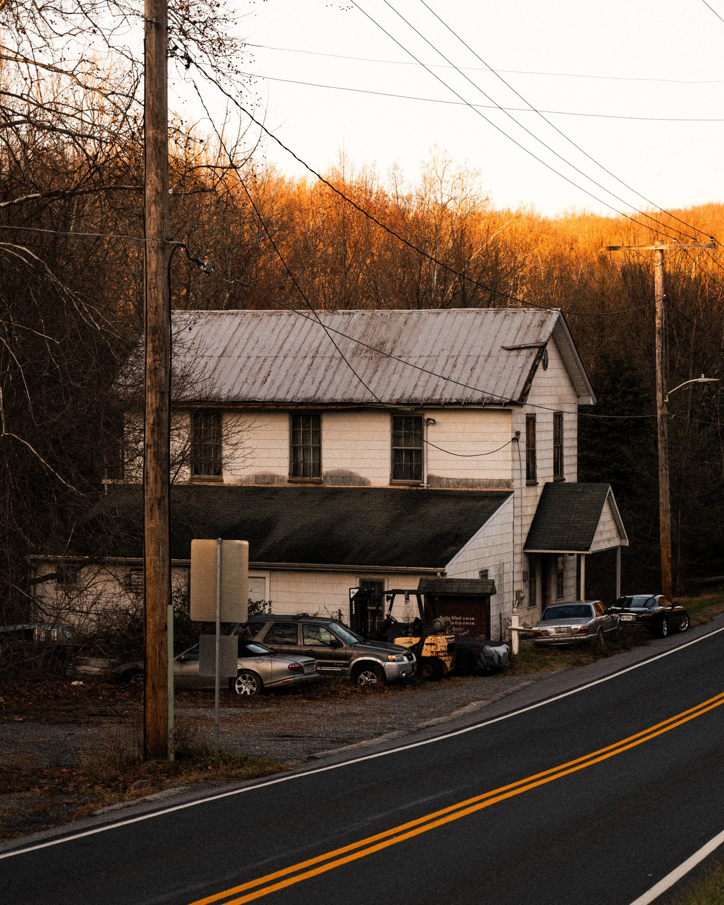 An old two-story house with a metal roof, surrounded by trees with autumn colors. There are multiple cars parked in front and a forklift next to the house. Power lines run above the scene and a road with double yellow lines is in the foreground.