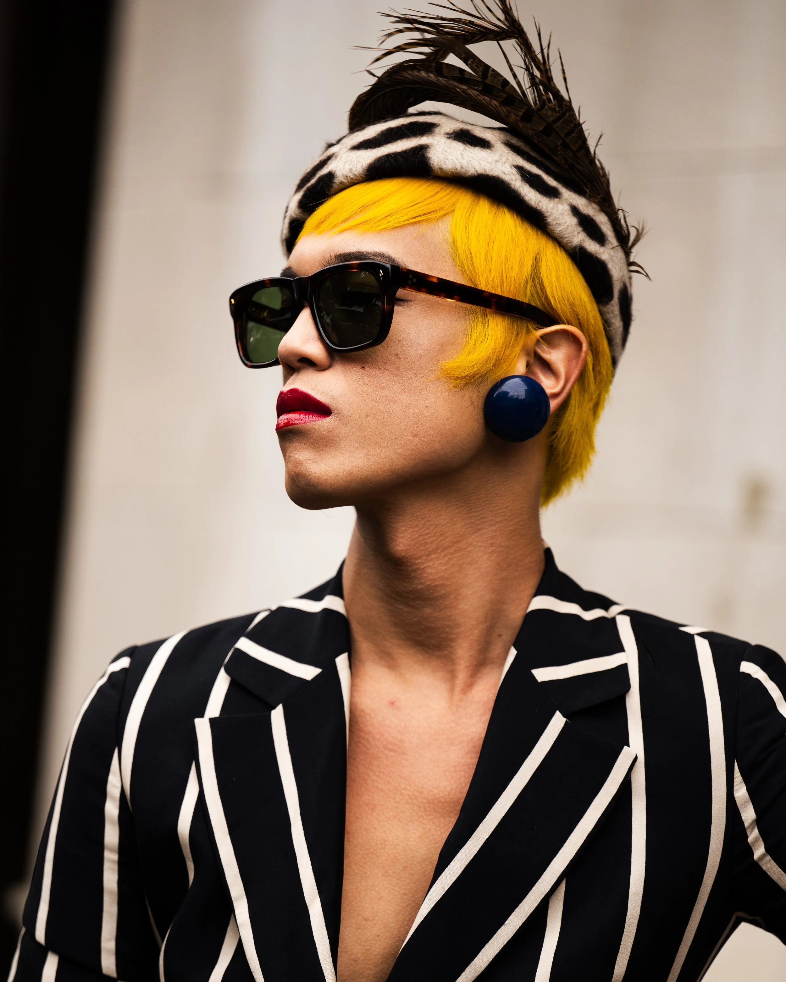 A woman with bright yellow hair, wearing black sunglasses, a leopard print headscarf with feathers, large dark earrings, red lipstick, and a black and white striped blazer.