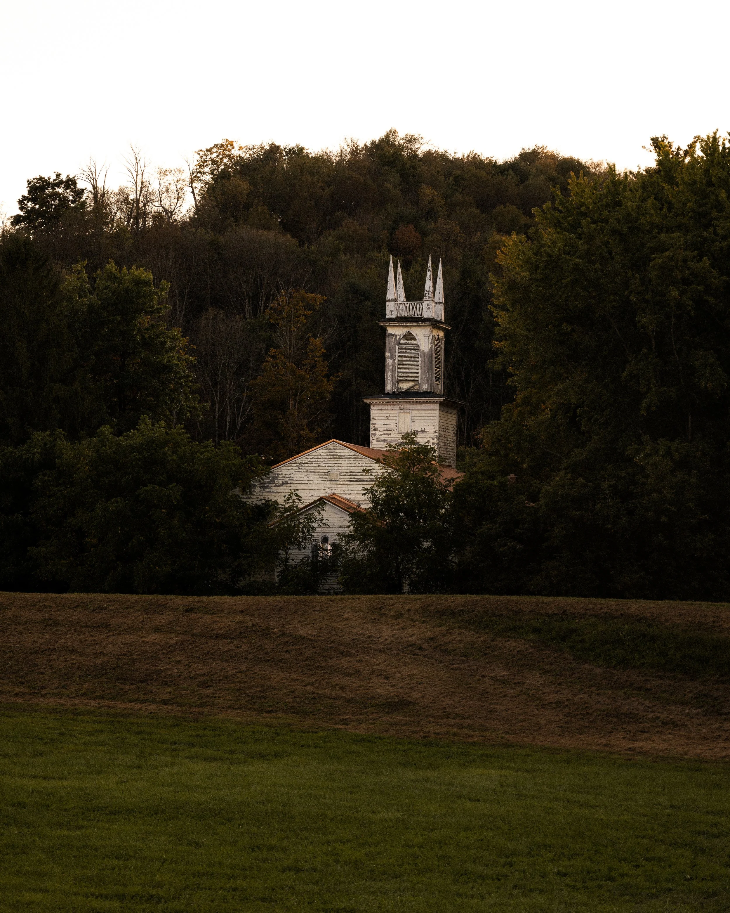 An old white church with a tall, narrow bell tower featuring pointed spires, surrounded by dense green trees on a hillside with a grassy foreground.