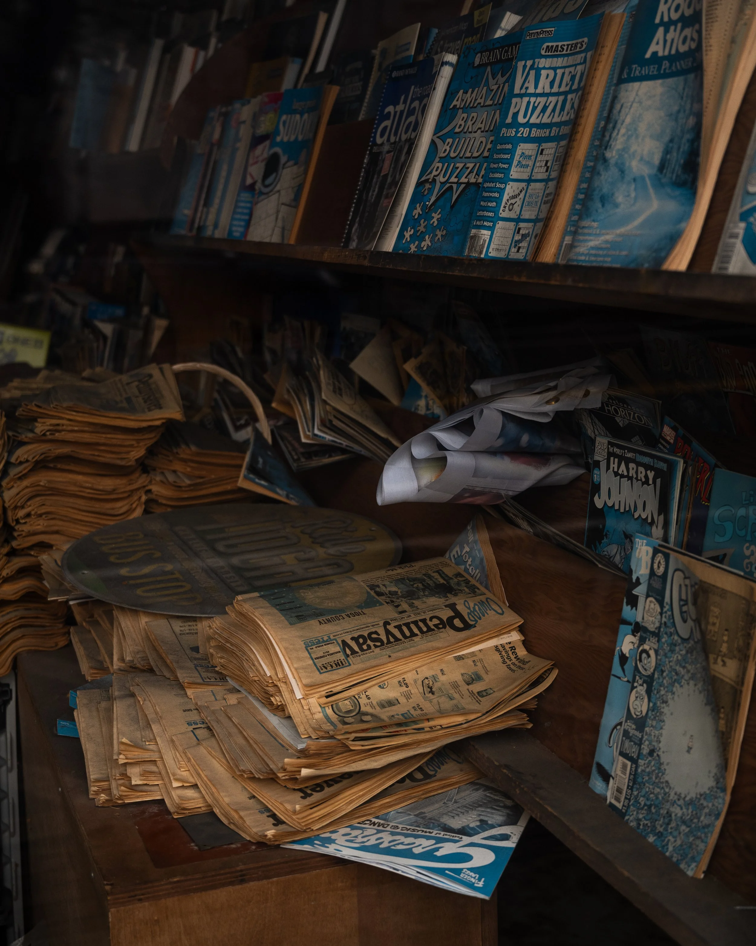 A cluttered wooden desk with newspapers, magazines, and paper scattered on top. Shelves above contain books and magazines, including titles about puzzles and comics.