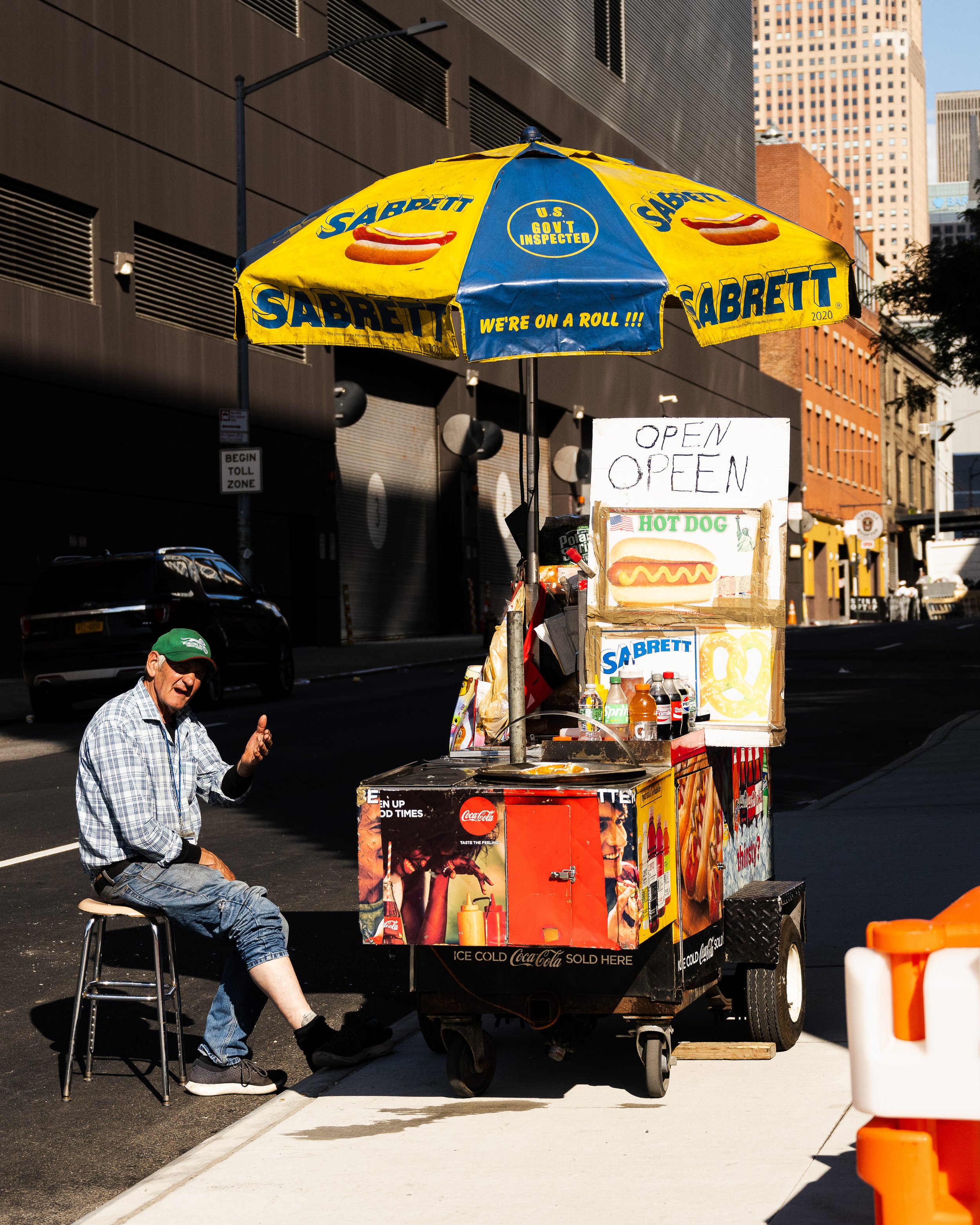 Street hot dog stand with yellow umbrella, signs that say 'OPEN' and 'HOT DOG,' and various condiments and drinks, on city street with man sitting nearby.
