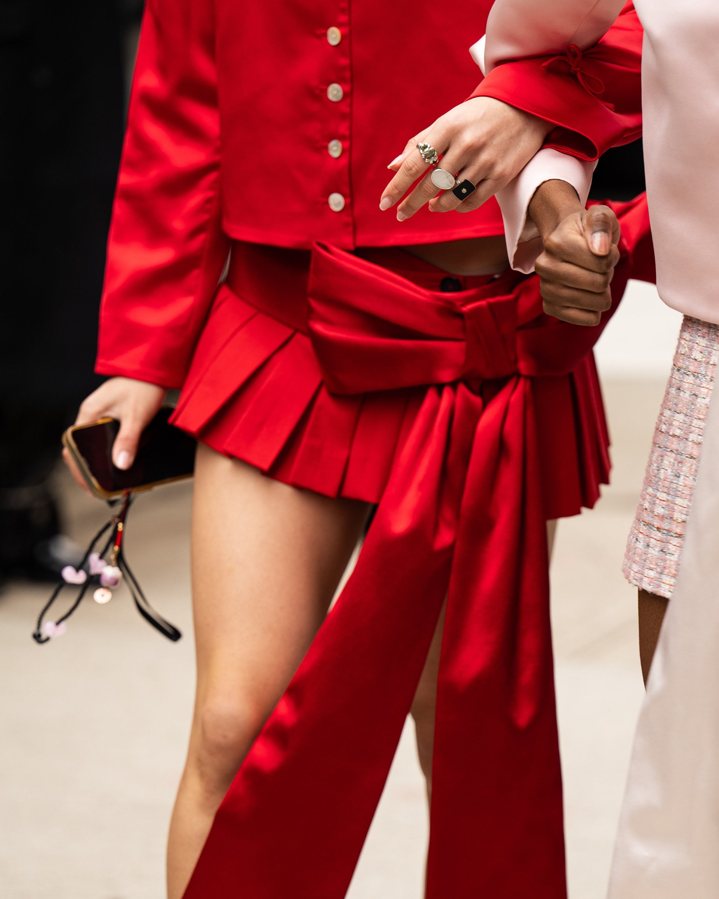 Two women, one in a red blouse and pleated red skirt, and another in a light pink blazer and skirt, holding hands. Street style photograph by street style photographer Kyle Mark Peterson during NYFW.