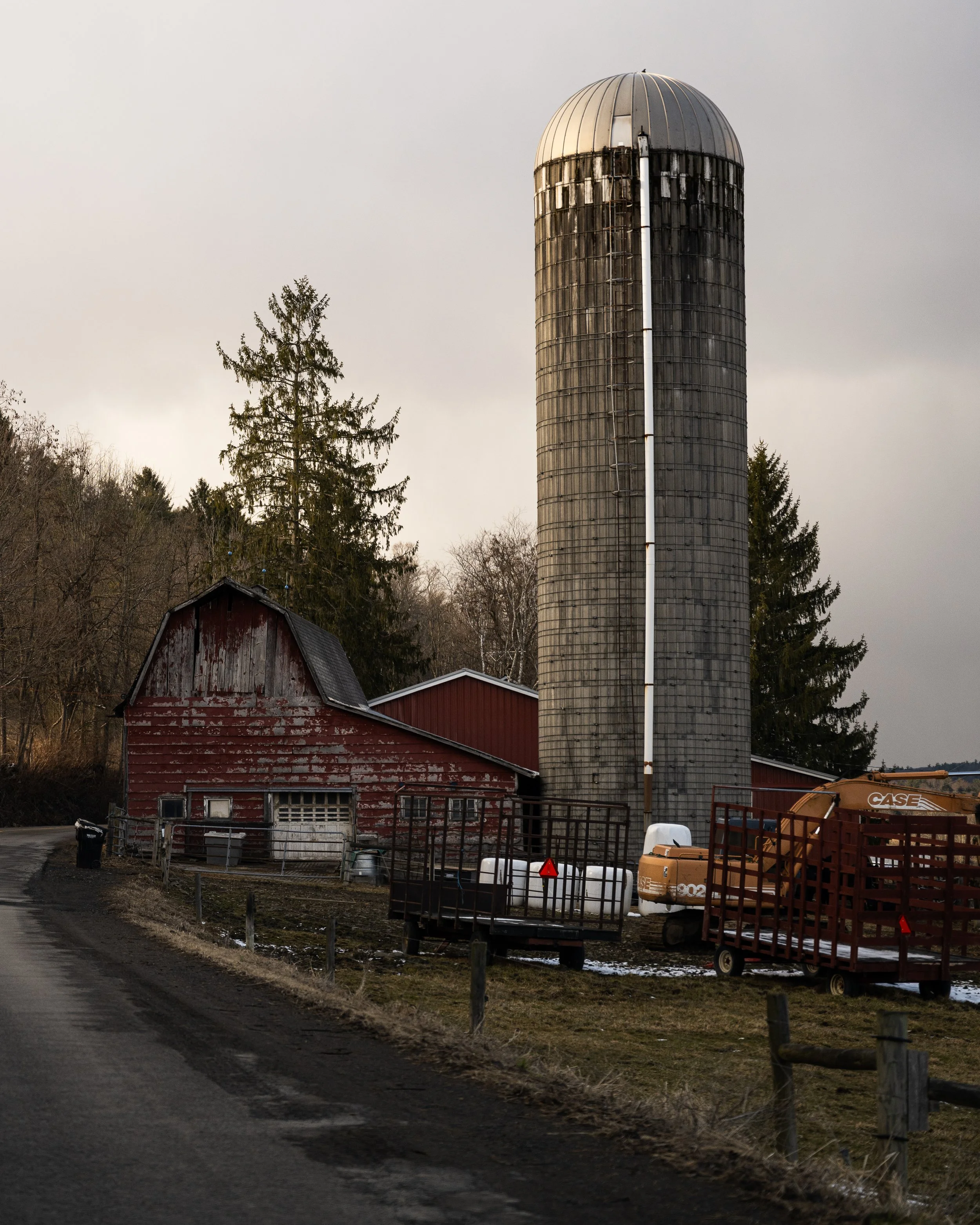 A rural scene with an old red barn, a tall gray silo, and farm equipment on a dirt path, with trees and overcast sky in the background.
