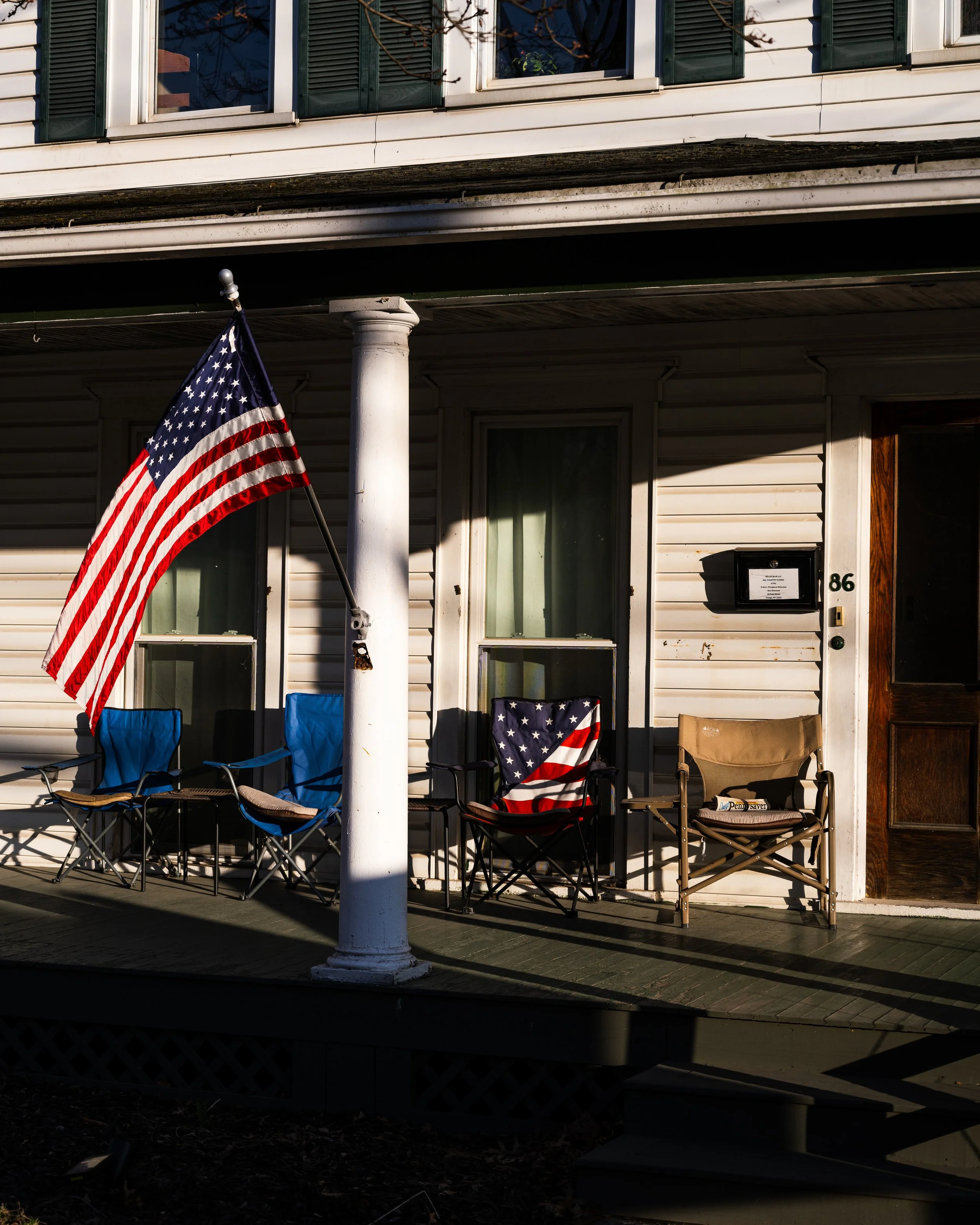 Front porch of a house with an American flag, two blue chairs, a red, white, and blue chair cover, and a beige chair, with house number 86 and a mailbox.