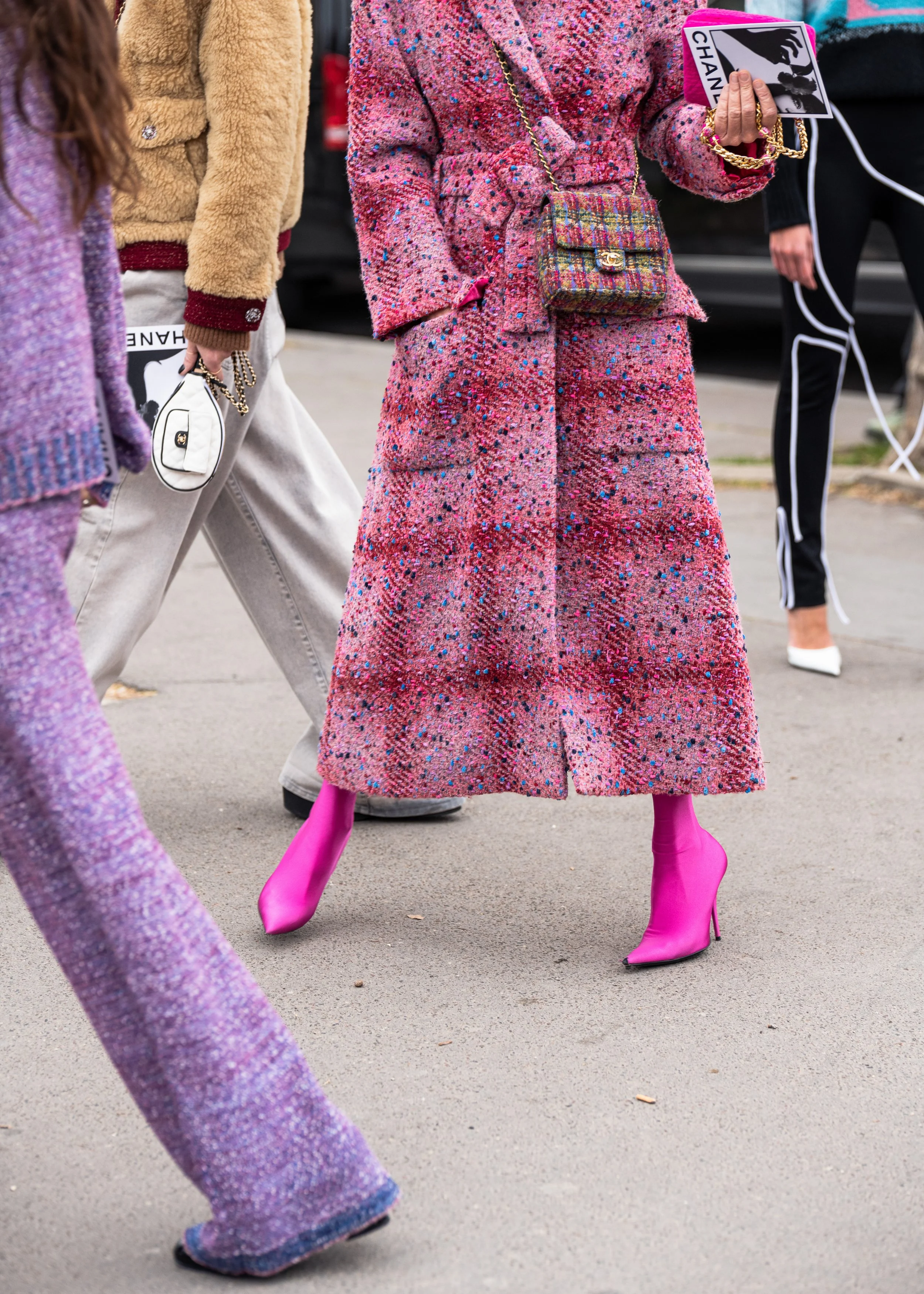 Person wearing a long pink, red, and purple textured coat with bright pink stiletto boots, holding a small multicolored purse and a magazine. Street style photograph by street style photographer Kyle Mark Peterson during Paris Fashion Week.
