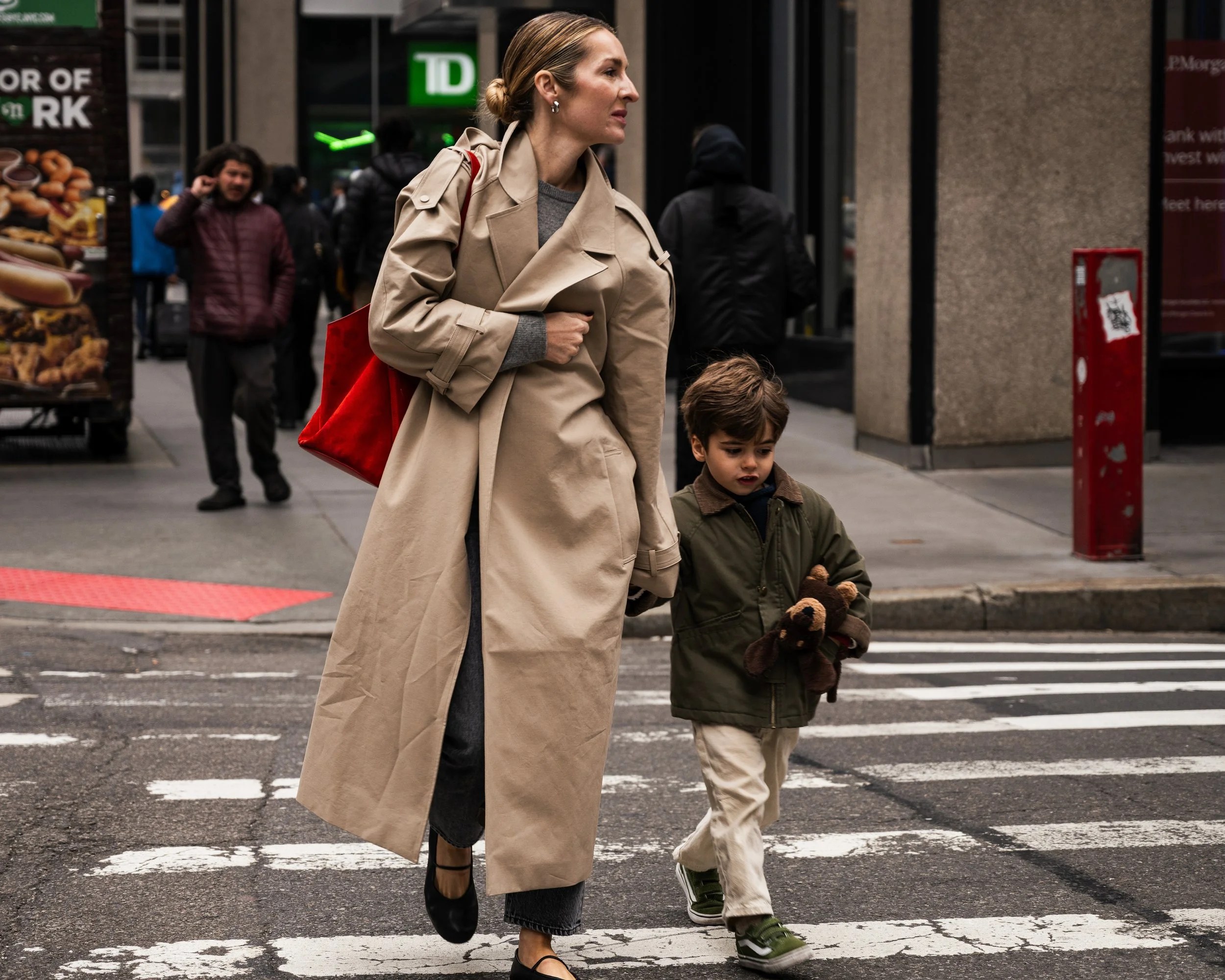 A woman and a young boy crossing the street in an urban area, with various people and storefronts in the background.
