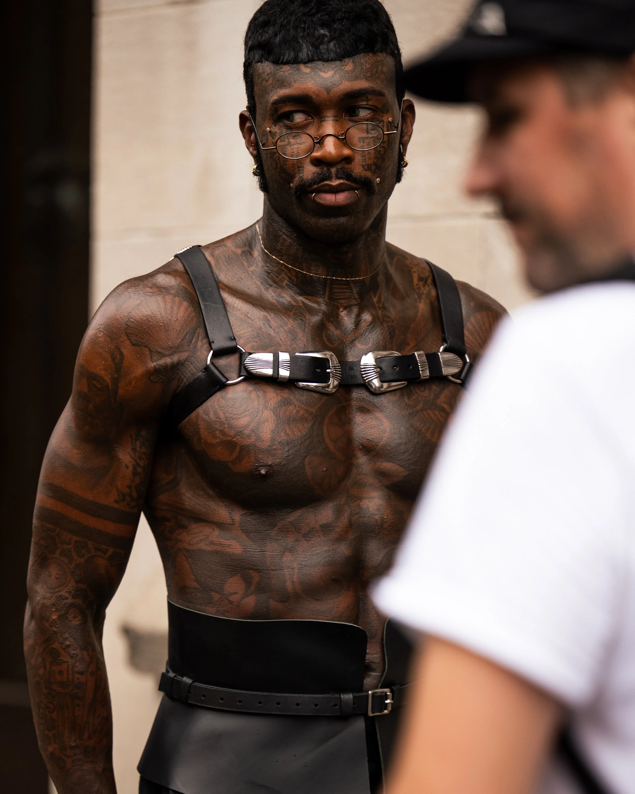 A tattooed man with glasses and earrings looking to the side, wearing a harness and black pants, while conversing with a blurred person in the foreground. New York Fashion Week street style imagery by Kyle Mark Peterson.