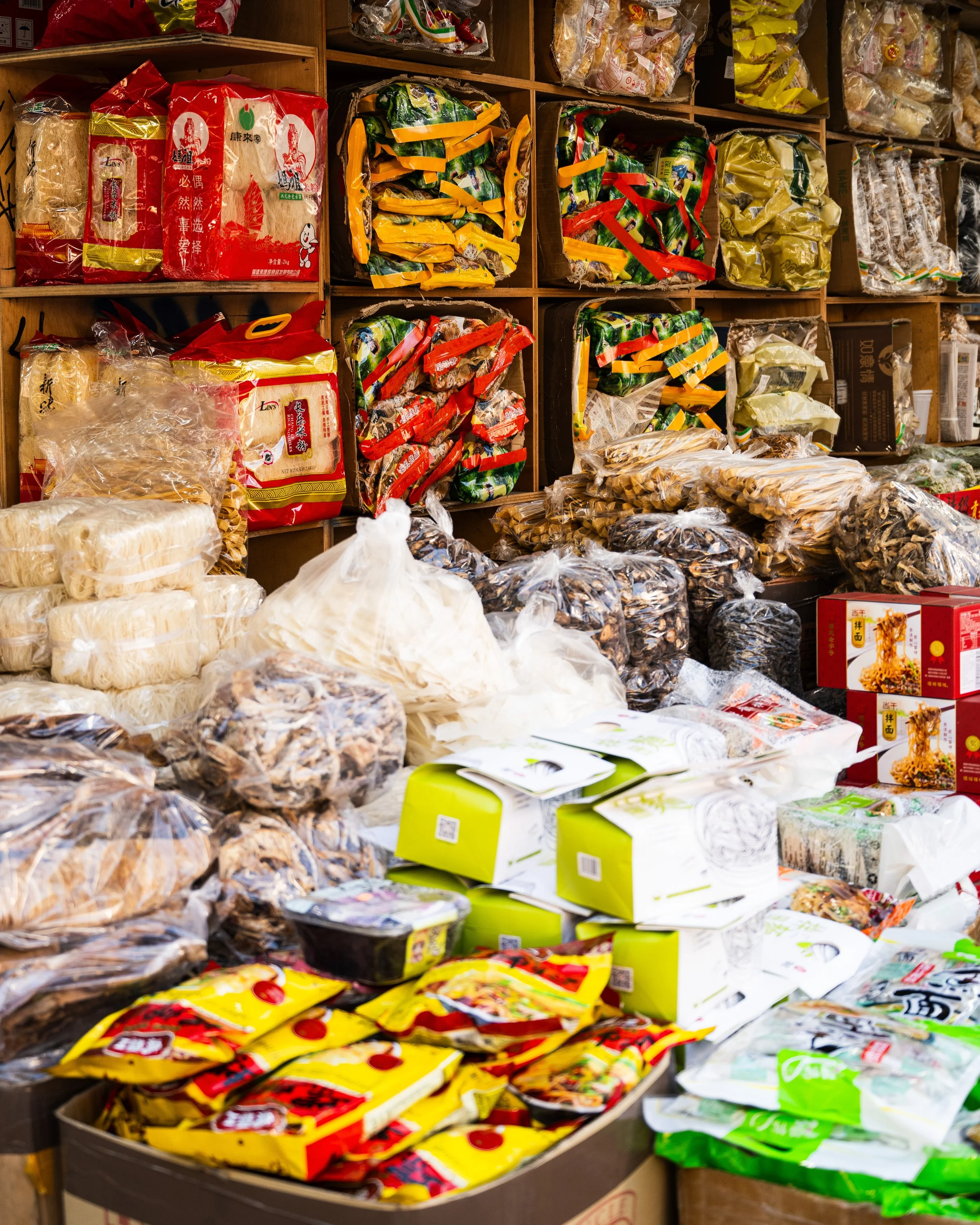 Shelves and tables filled with various packaged Asian food products, including noodles, dried mushrooms, and snack packets.
