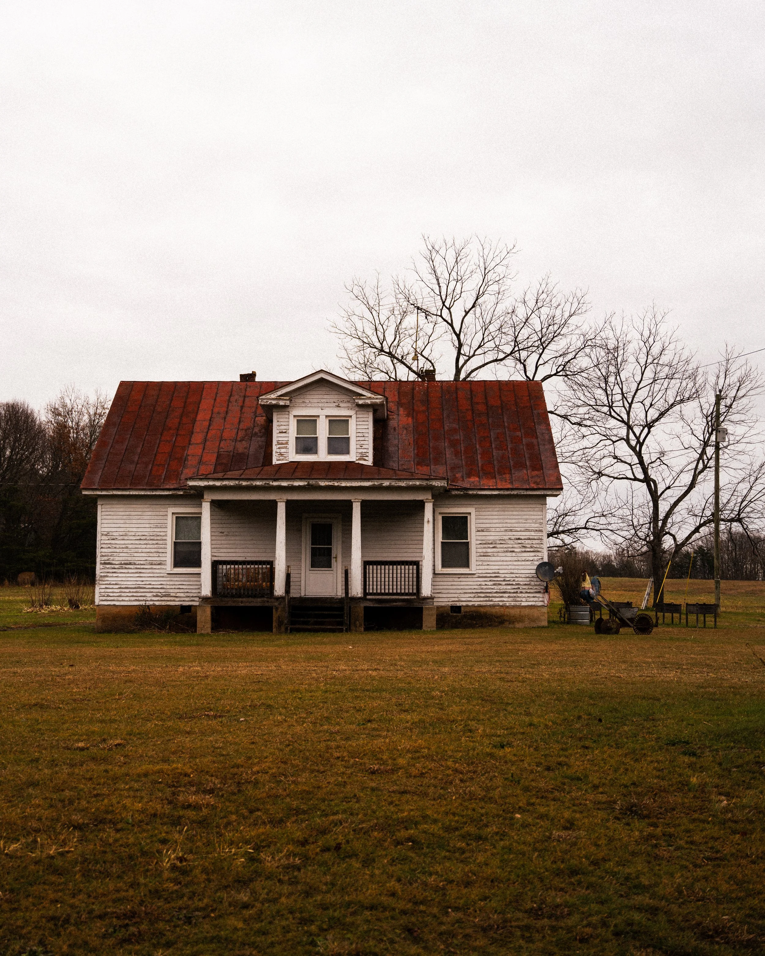 Old white wooden house with a red rusted metal roof, small front porch, and leafless trees in the background, on a grassy field under an overcast sky.