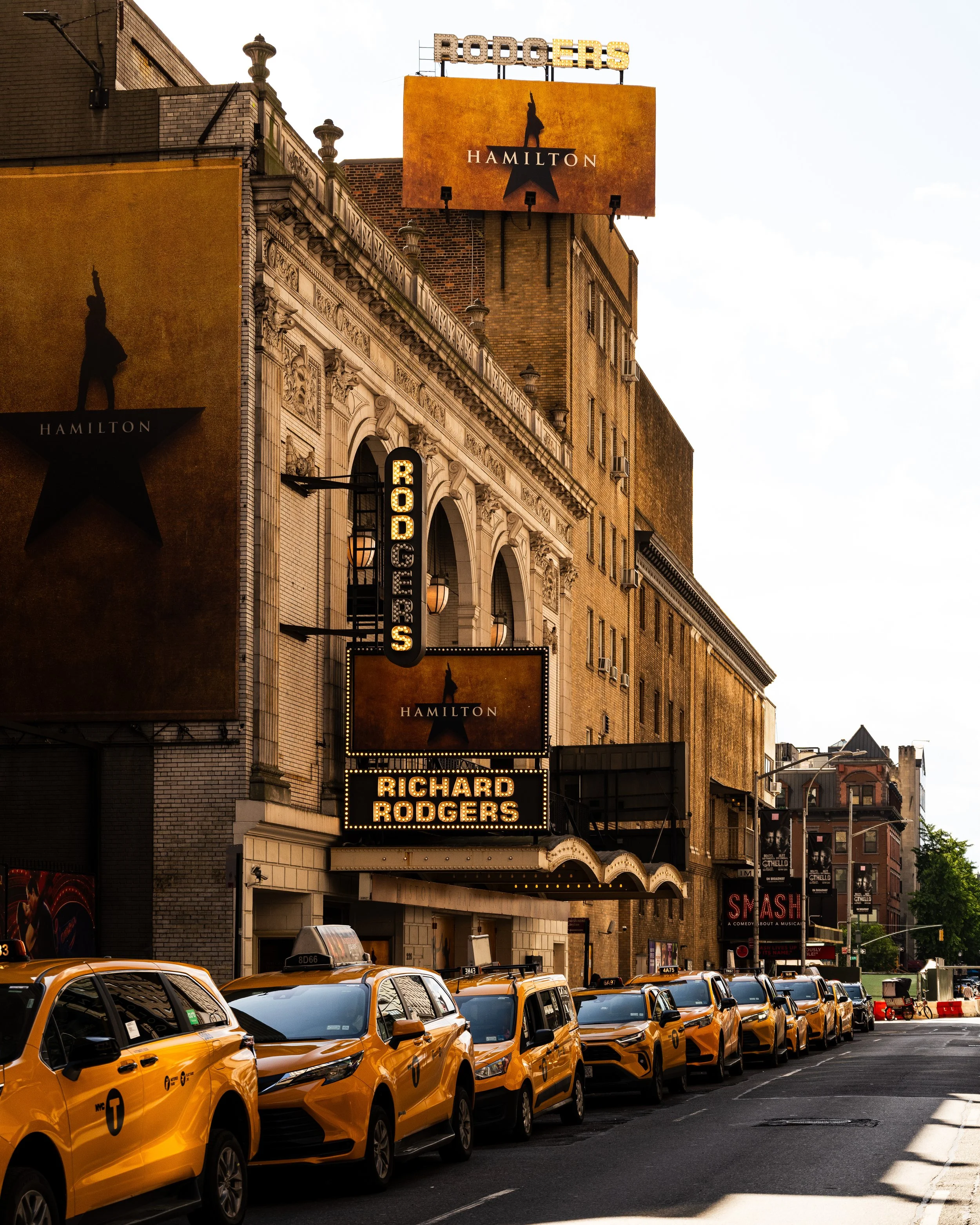 Line of yellow taxis parked outside Richard Rodgers Theatre in New York City, with marquee signage for the musical 'Hamilton' and posters for 'Smash' on the building.