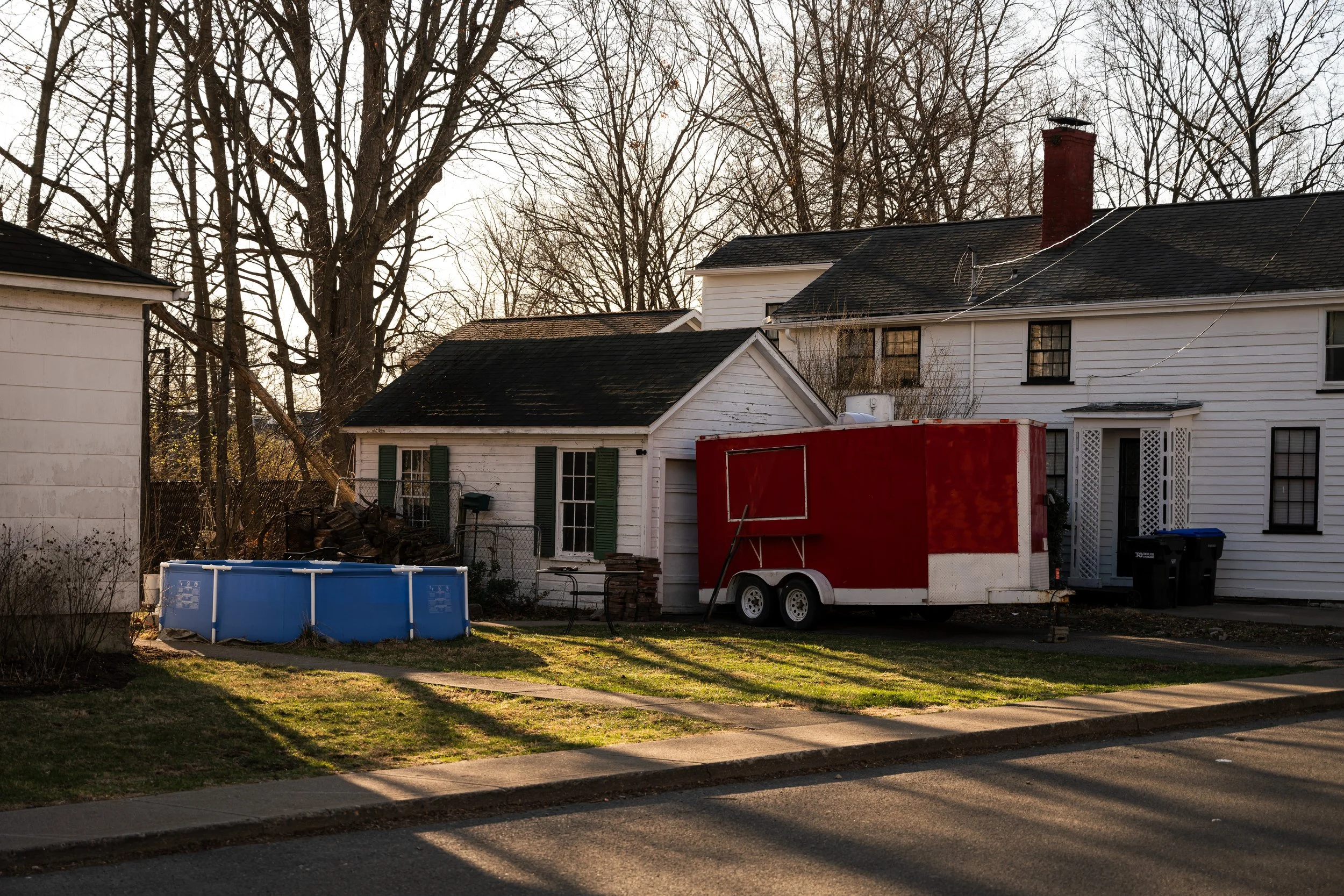 Residential backyard with a red food trailer, a blue pool, and a small white house with green shutters, surrounded by bare trees.
