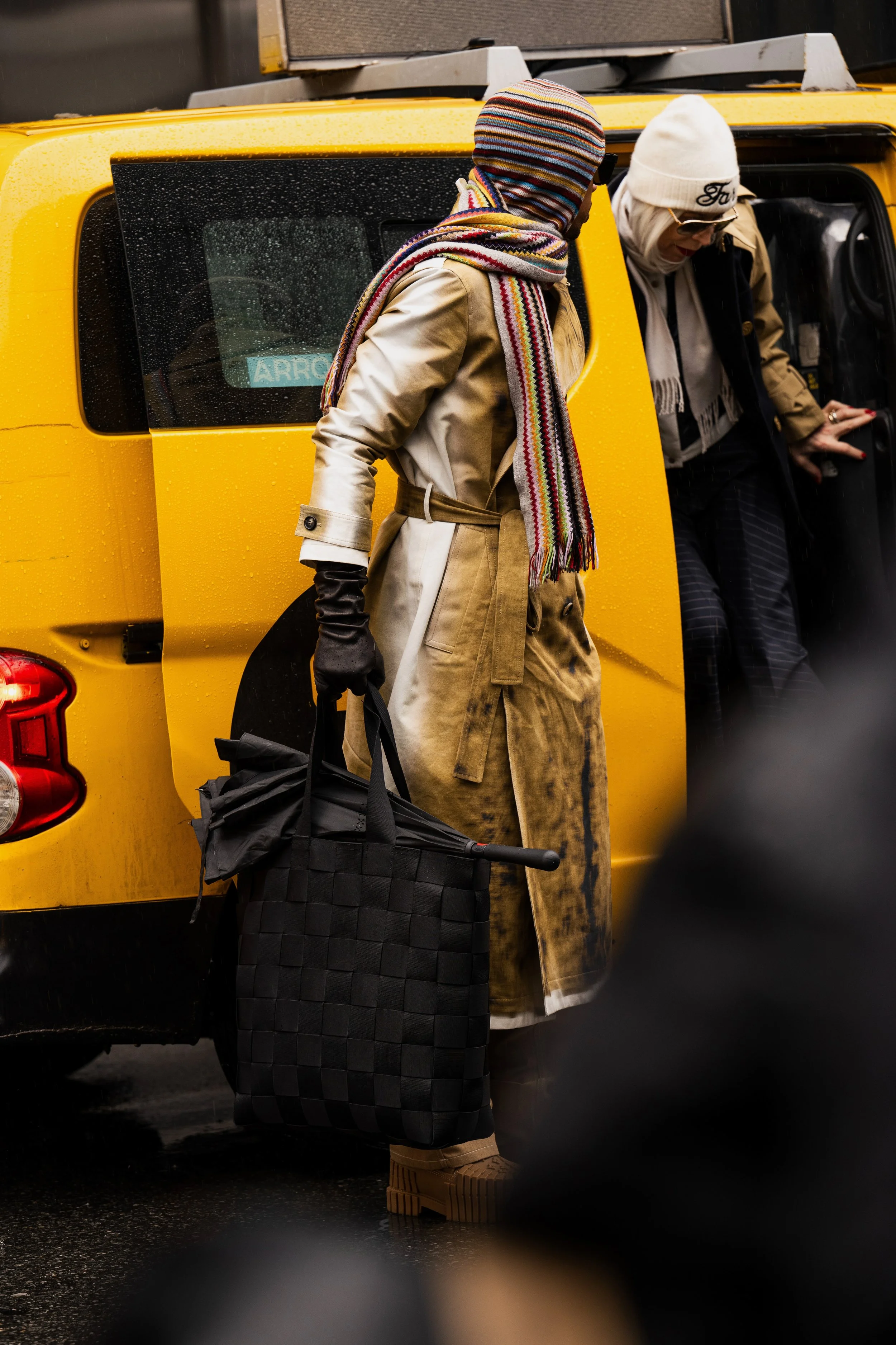 A woman dressed in a beige trench coat, striped hat, and scarf, carrying a large black handbag, stands near the open door of a yellow taxi on a rainy day.