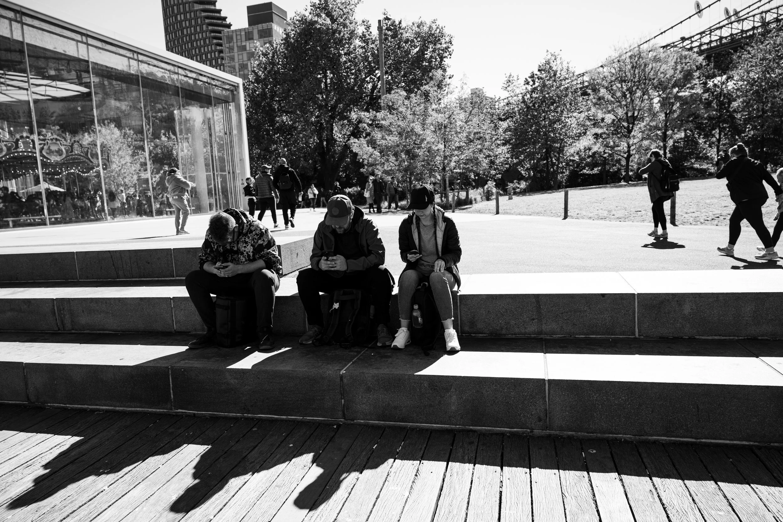 Three people sitting on a bench checking their phones in a park, with a glass building and people walking in the background in black and white.