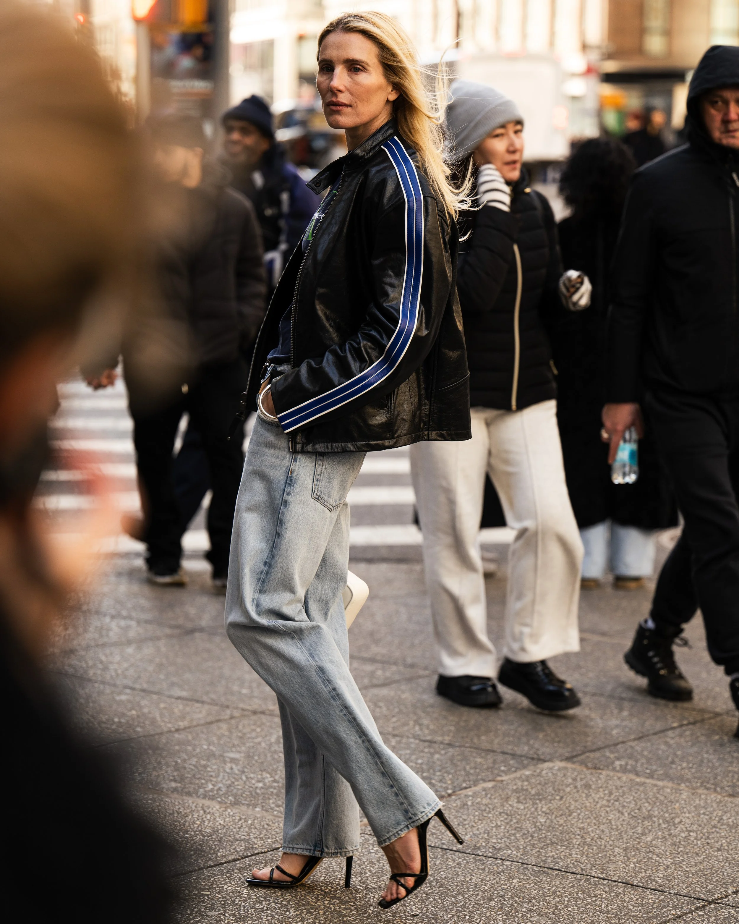 A woman with long blonde hair wearing a black leather jacket with blue stripes, light blue jeans, and black high heels, stands on a city crosswalk among pedestrians during daytime.