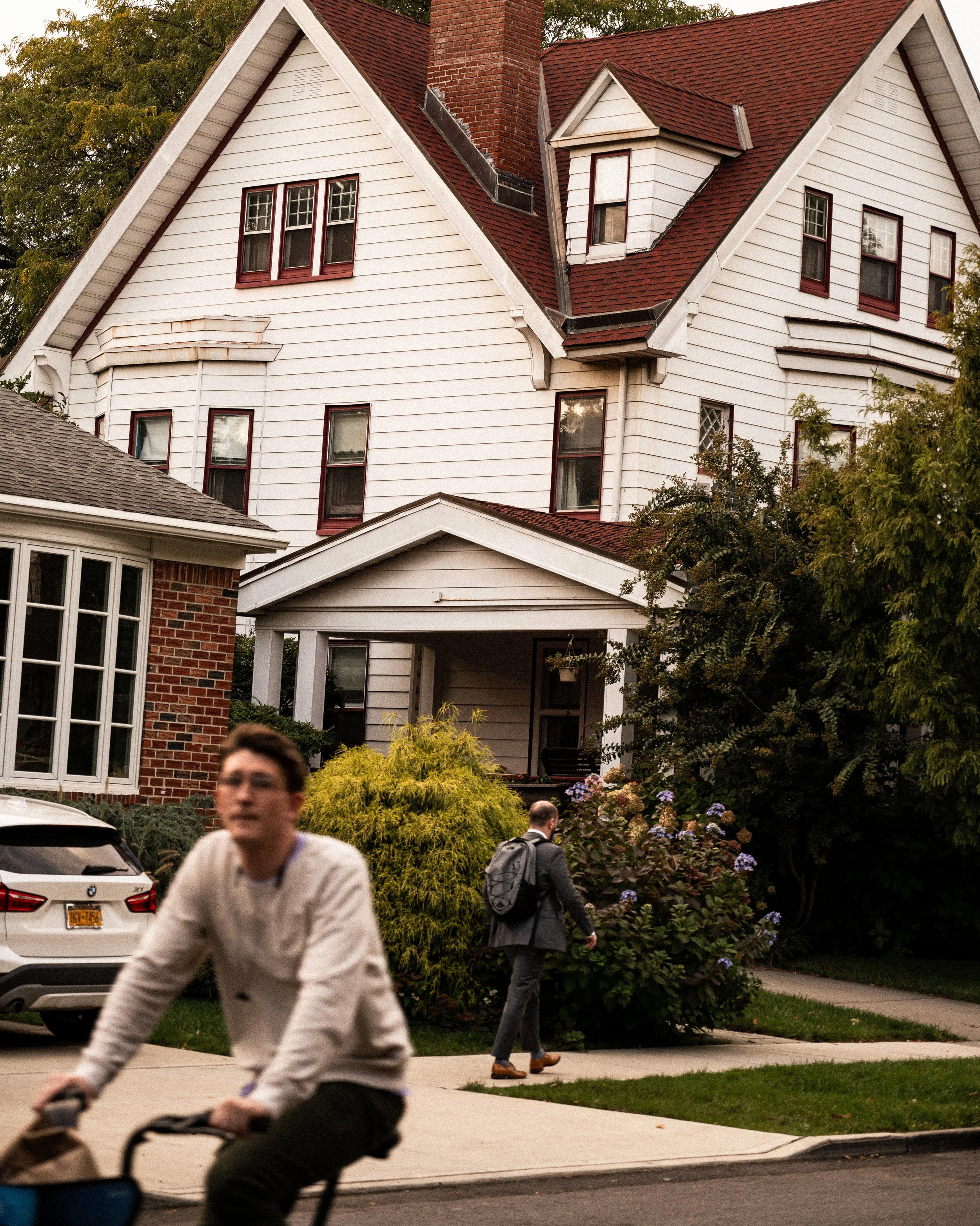 A residential neighborhood scene showing a white multi-story house with red window frames and a red roof, a person riding a bike in the foreground, and another person with a backpack walking on the sidewalk.