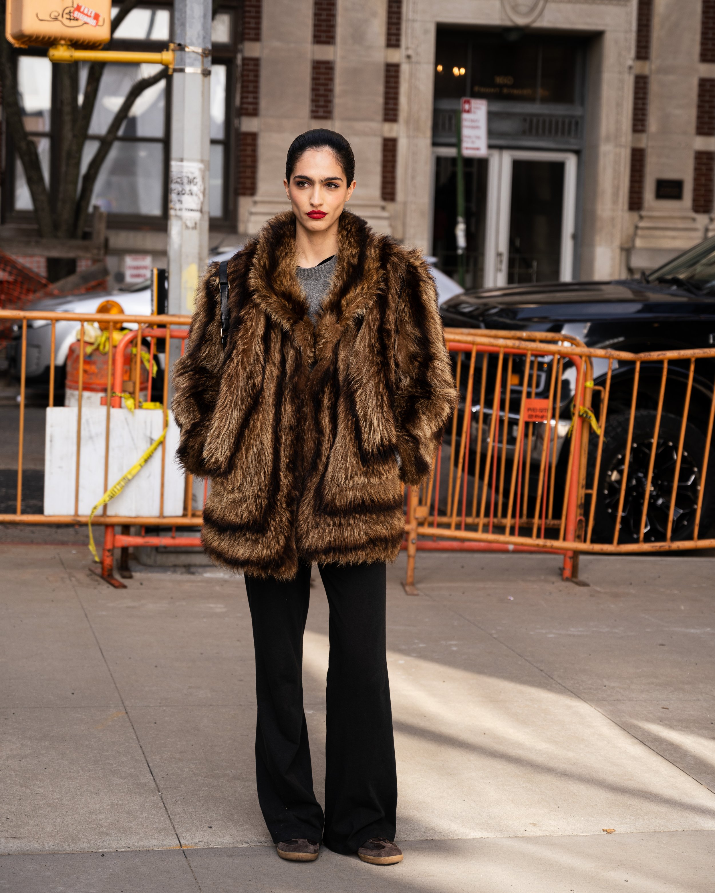A woman wearing a large fur coat standing on a city sidewalk. Street style photograph by street style photographer Kyle Mark Peterson during NYFW.