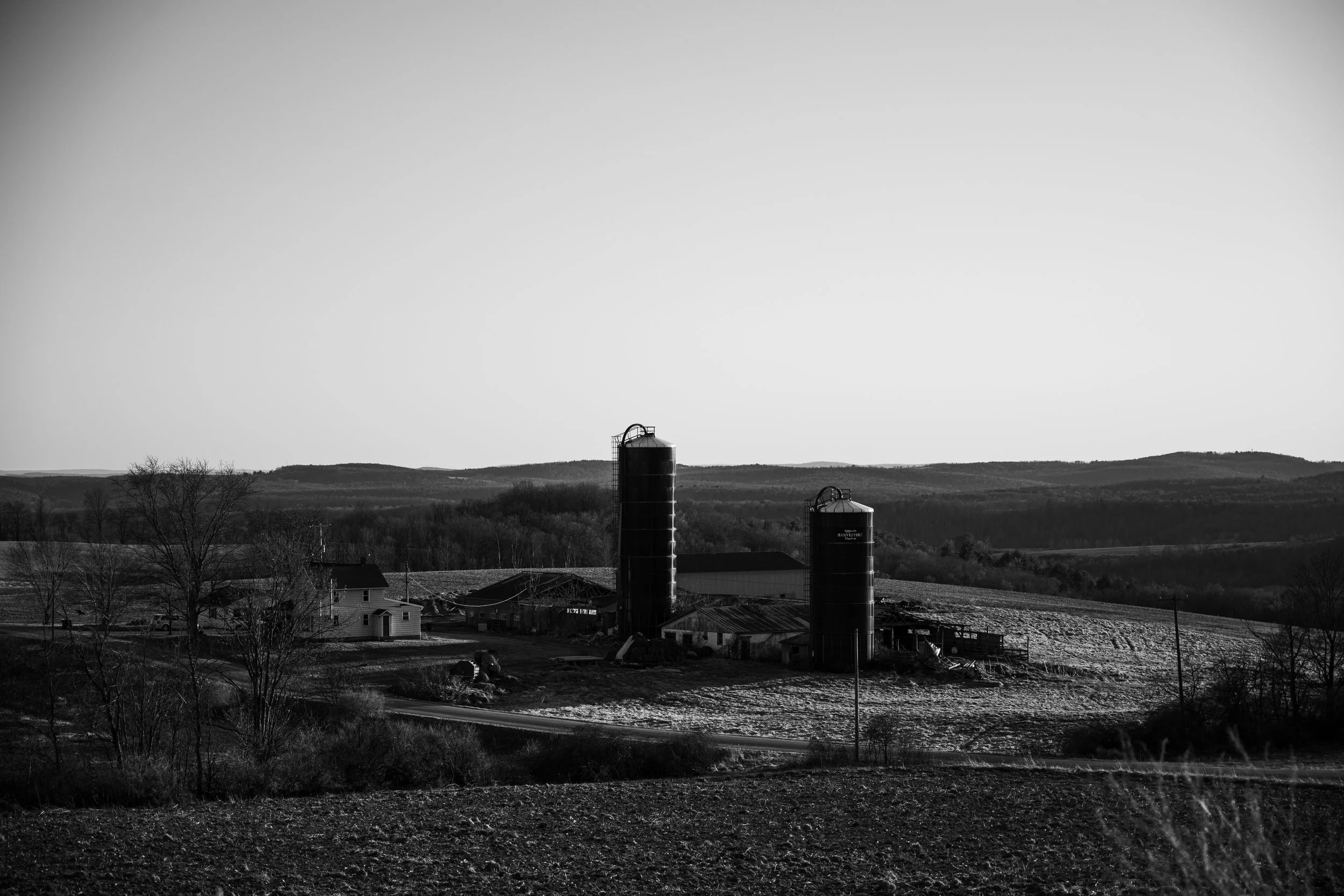 Black and white photo of a rural farm landscape with two large silos, a farmhouse, and smaller farm buildings, with rolling hills in the background.