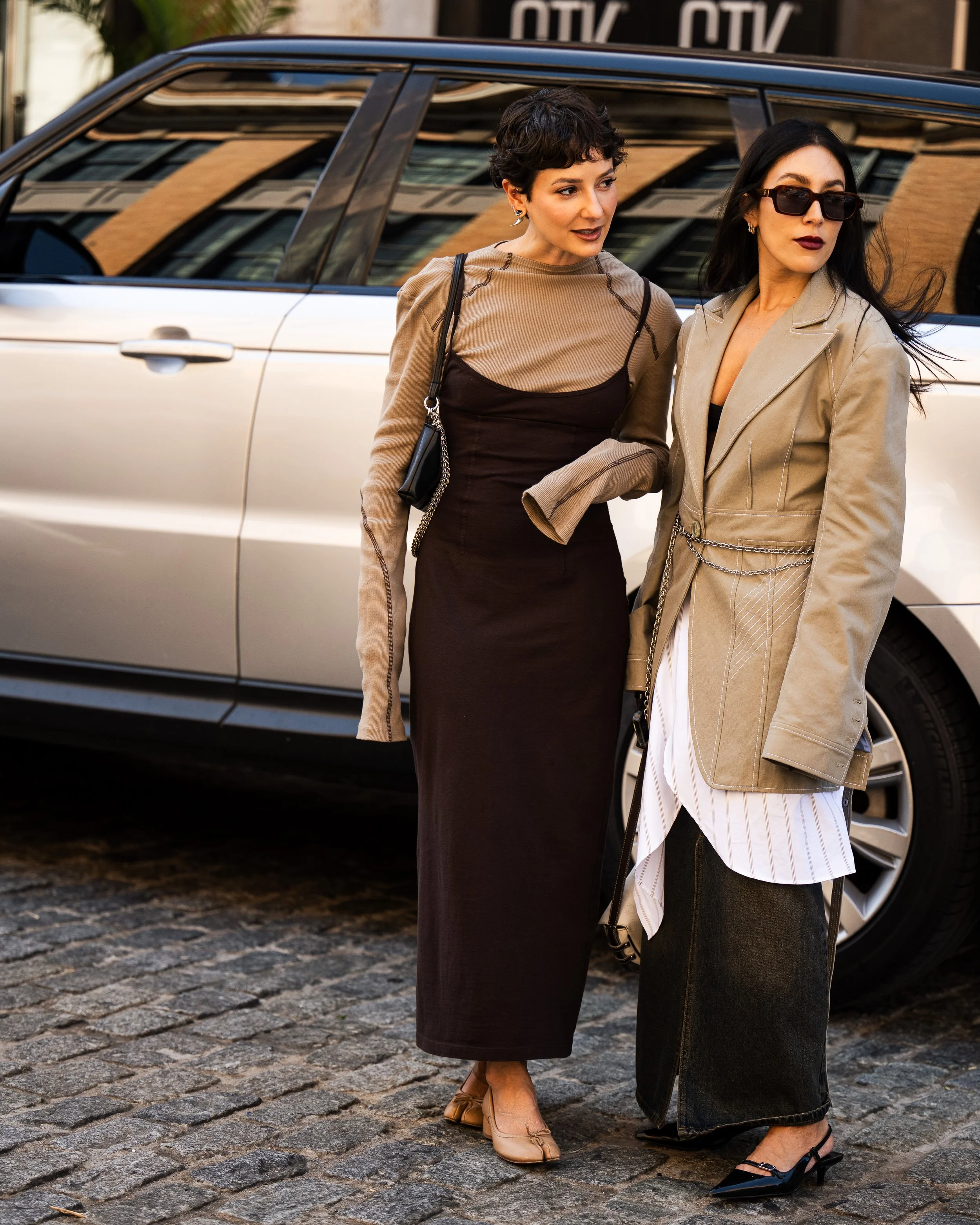 Two women standing on a cobblestone street in front of a silver car, engaged in conversation. They are dressed stylishly, one wearing a long black dress with a sheer overlay and the other wearing a beige blazer with dark sunglasses.