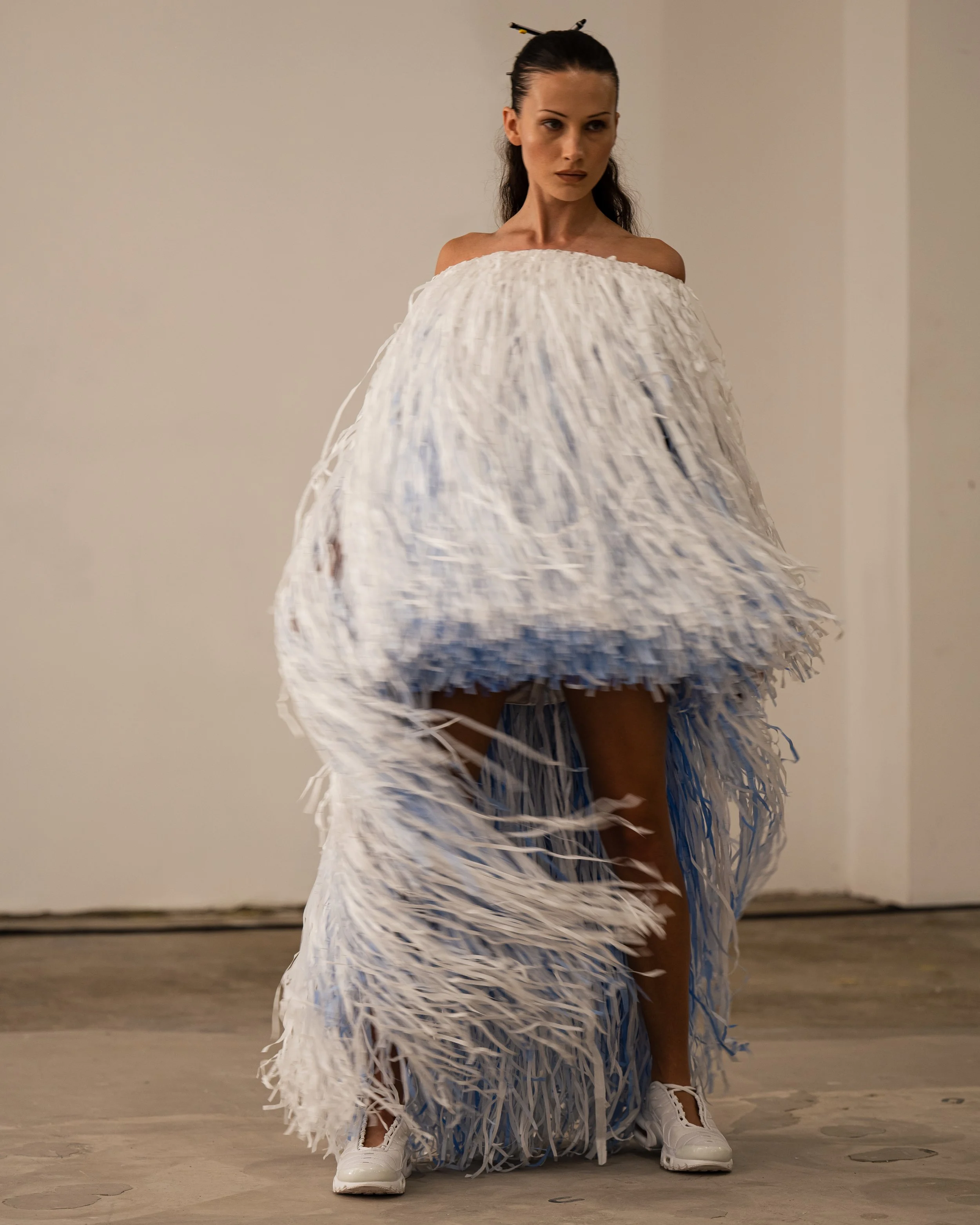Model in a white, fringed, layered dress with a high slit, wearing white sneakers, standing against a plain wall. Backstage photograph by fashion photographer Kyle Mark Peterson during NYFW.