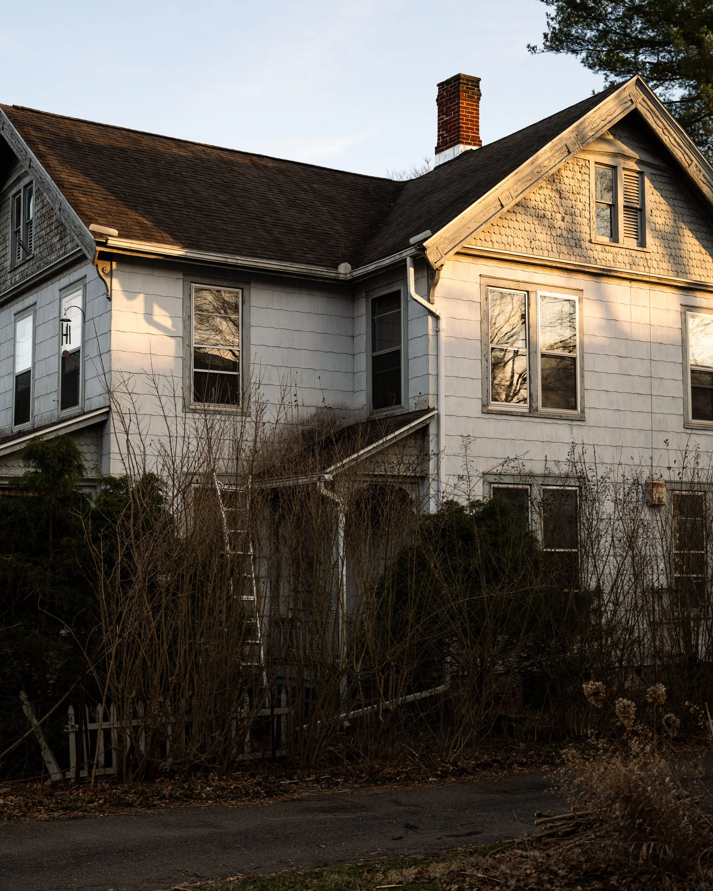 An old, white, two-story house with missing shingles, surrounded by overgrown bushes and a small cracked pathway in front.