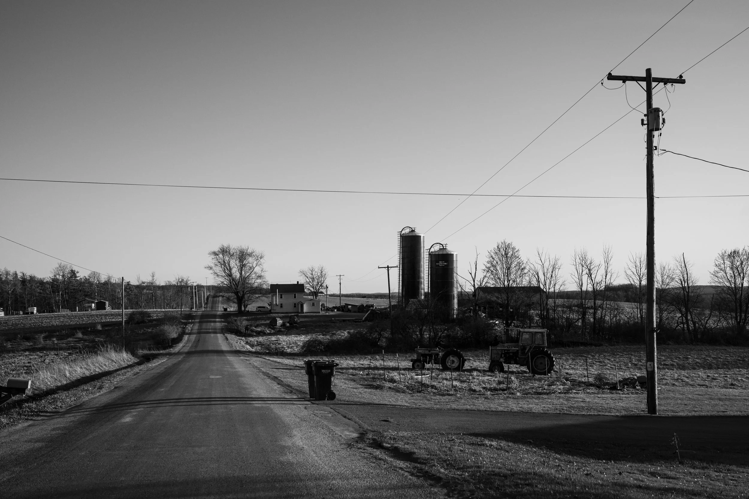 A rural farm scene in black and white with a dirt road, utility pole, barn, silos, and farming equipment, with leafless trees and a clear sky.