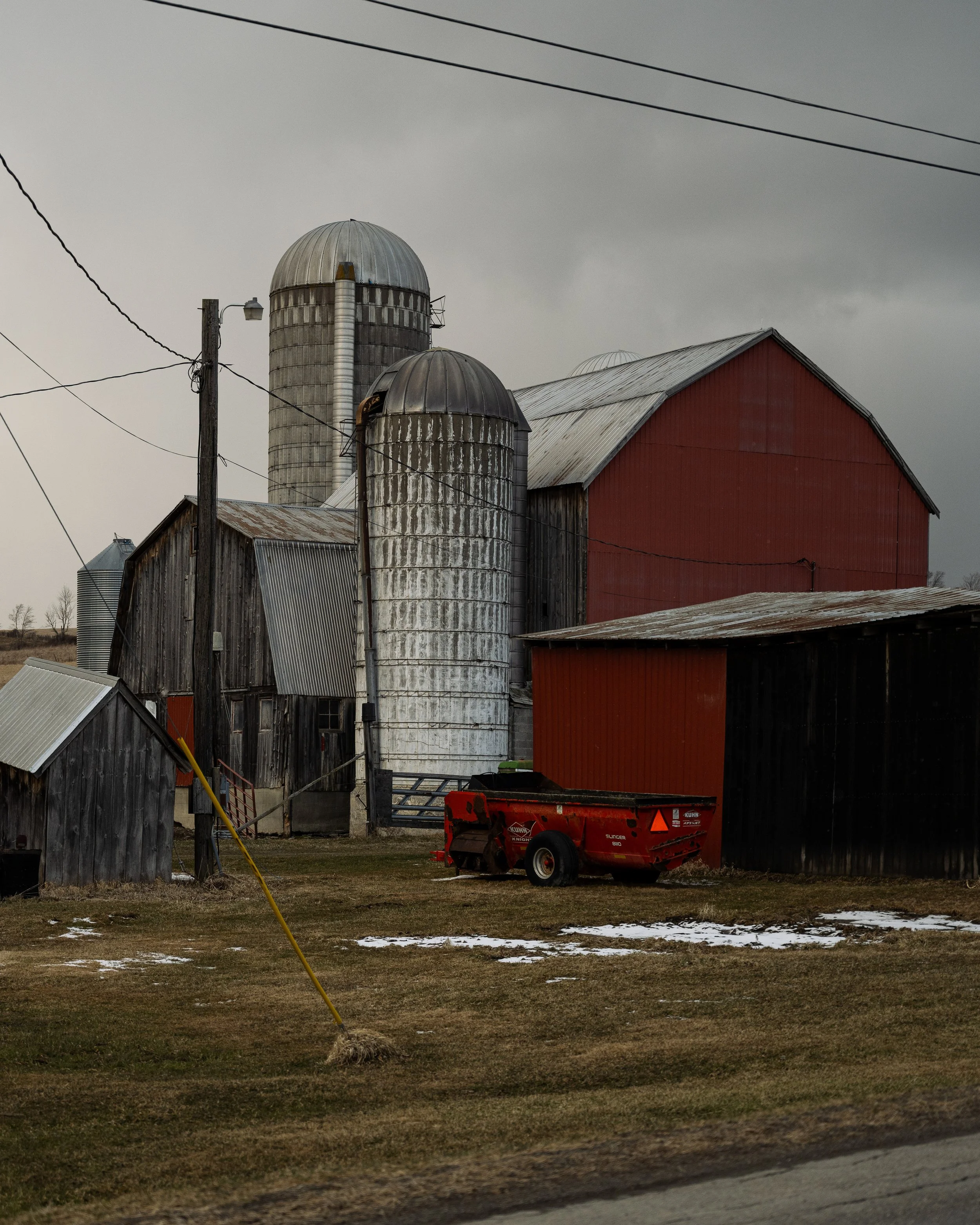 A farm scene with old barn buildings, silos, a red farm wagon, and overcast skies.