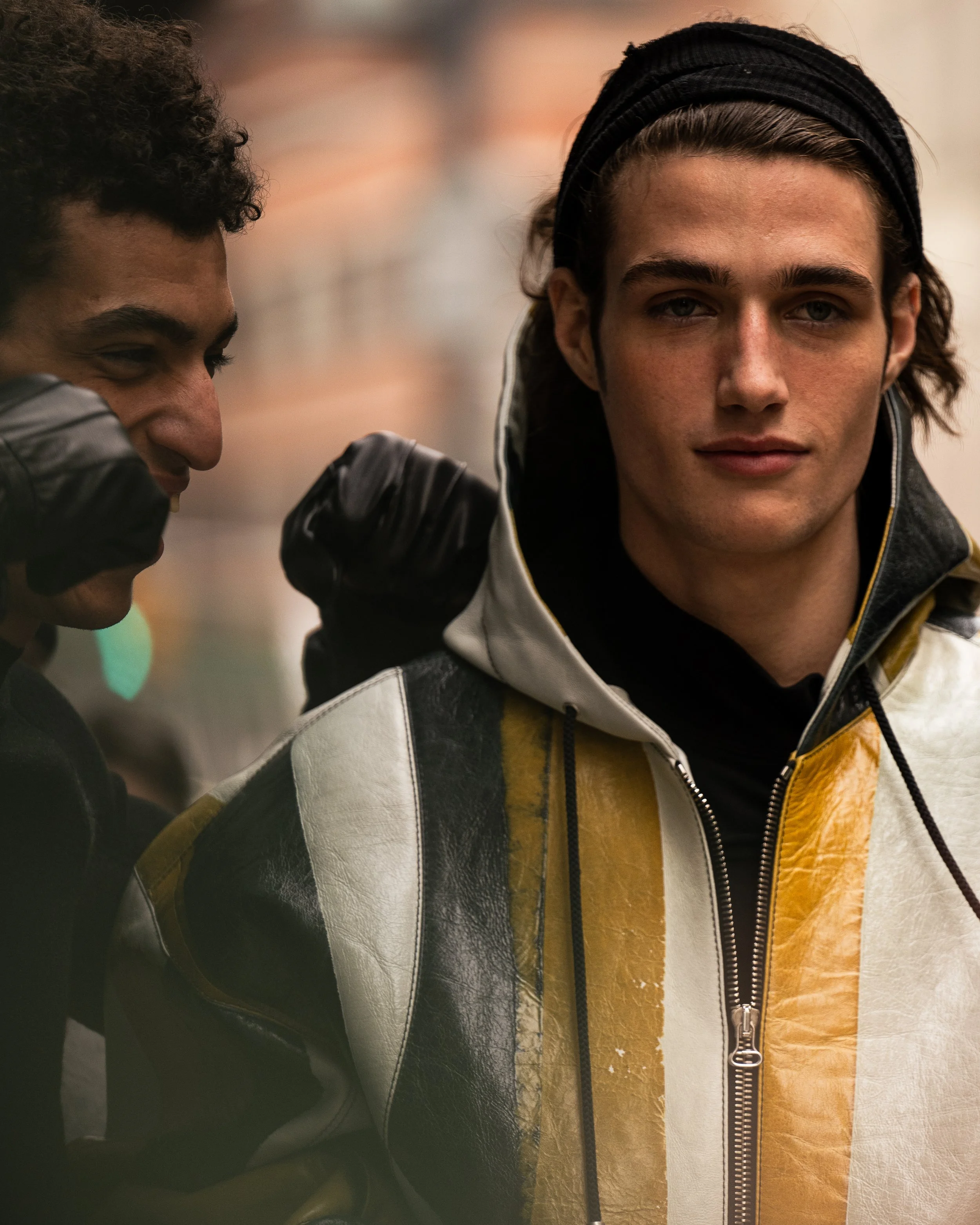 Two young men standing closely, one smiling with his eyes closed, the other looking directly at the camera with a neutral expression, in an urban setting. Street style photograph by street style photographer Kyle Mark Peterson during NYFW.