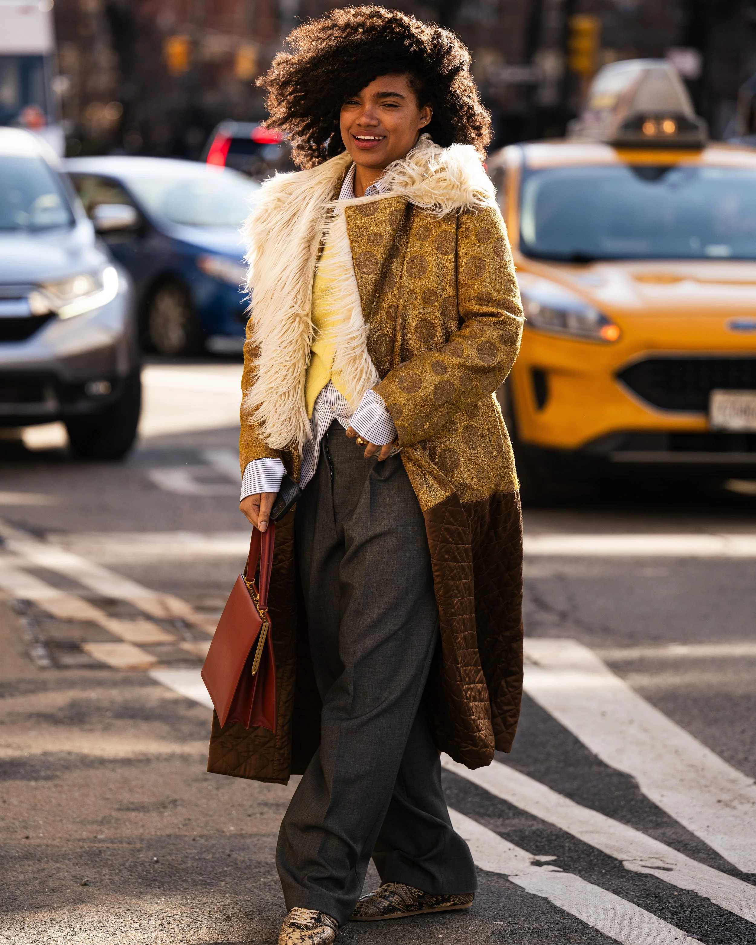 A woman with curly hair crossing a city street, dressed in a long coat, with a fur scarf, and holding a brown suitcase and red bag.