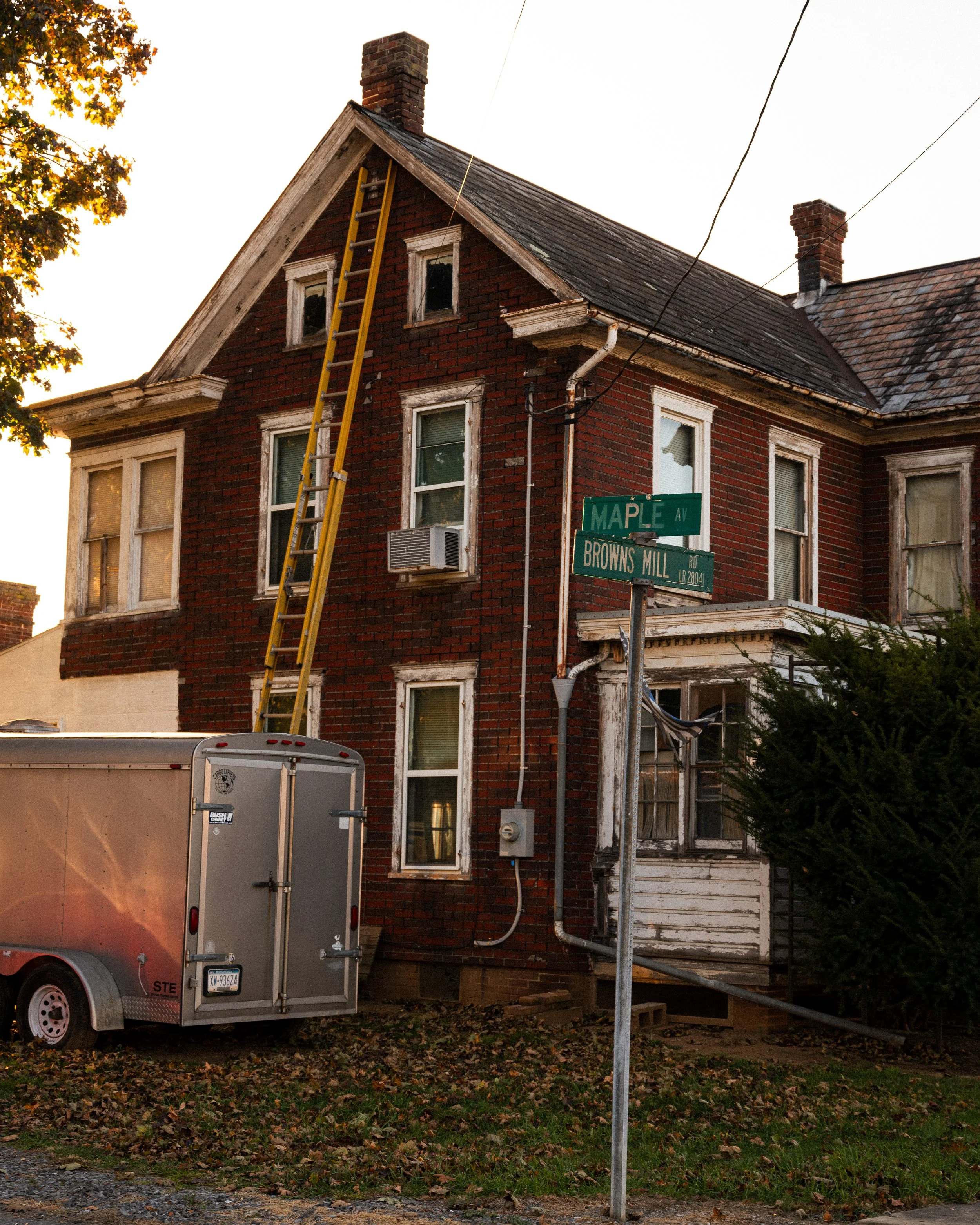 A two-story brick house with a pitched roof, multiple windows, and an air conditioning unit, along with a ladder leaning against it. The house appears to be undergoing maintenance or repair, with a trailer parked in front and street signs showing the intersection of Maple Ave and Browns Mill Rd.
