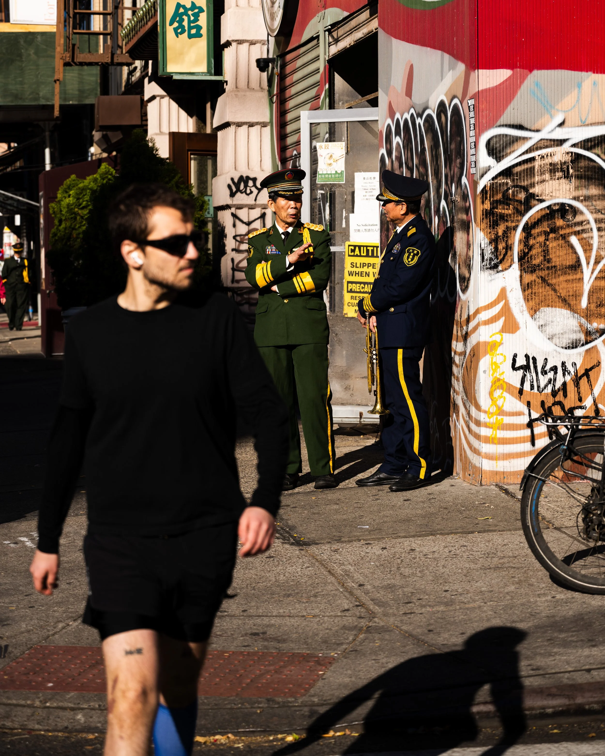 A man in black athletic clothes and sunglasses walking in the foreground, and two uniformed officers, one holding a trumpet, talking beside a graffiti-covered wall in an urban street scene.