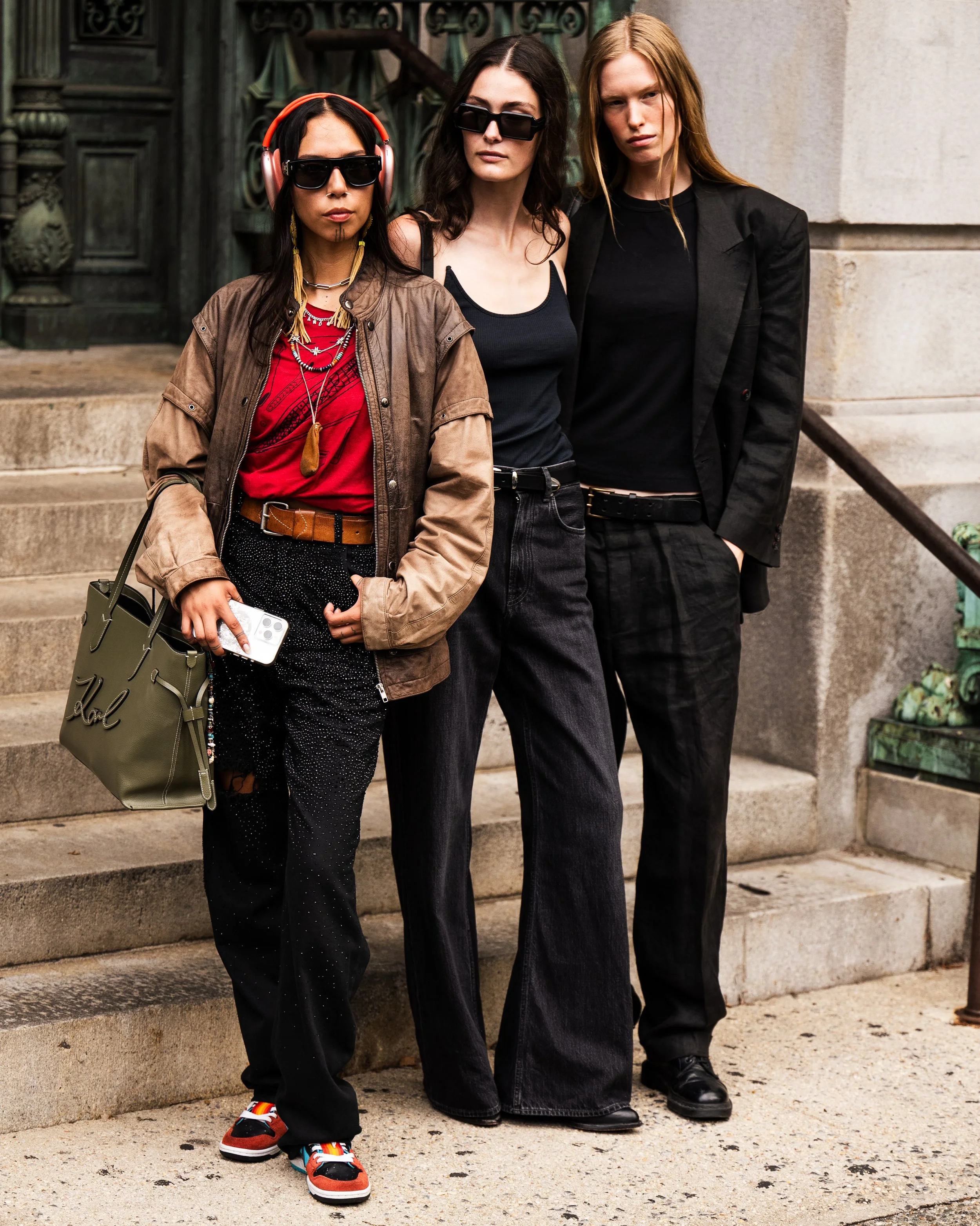 Three women standing on steps outside building, dressed in casual streetwear with sunglasses, one holding a smartphone and a tote bag. Street style imagery by Kyle Mark Peterson during New York Fashion Week.