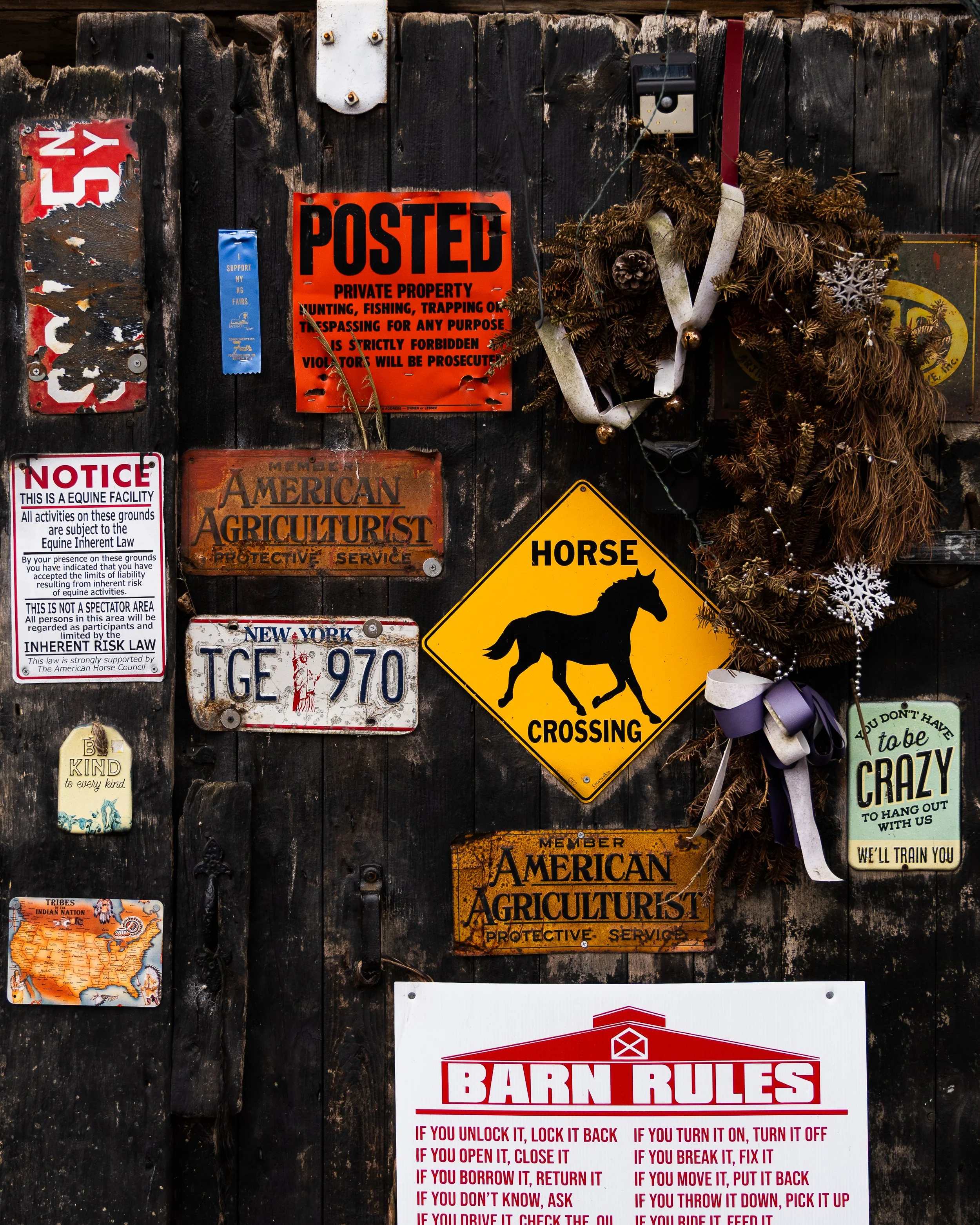 Black wooden wall decorated with various signs including a red 'POSTED' sign, a yellow 'Horse Crossing' sign, a white 'Notice' sign, a map of tribes in the United States, and a Christmas wreath with pinecones and ribbons.