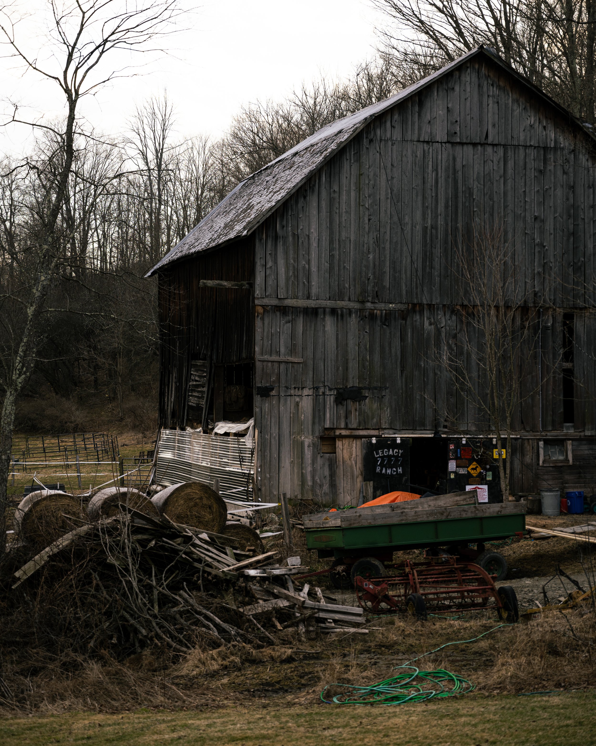 A rustic, weathered wooden barn in a rural setting with a leafless tree to the left and farm equipment, hay bales, and supplies scattered around the yard.