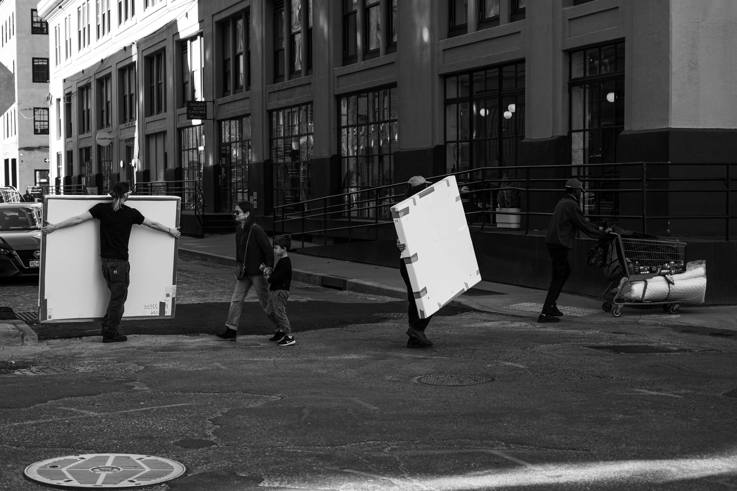 People moving across a city street, some carrying large boxes, with storefronts and buildings in the background.